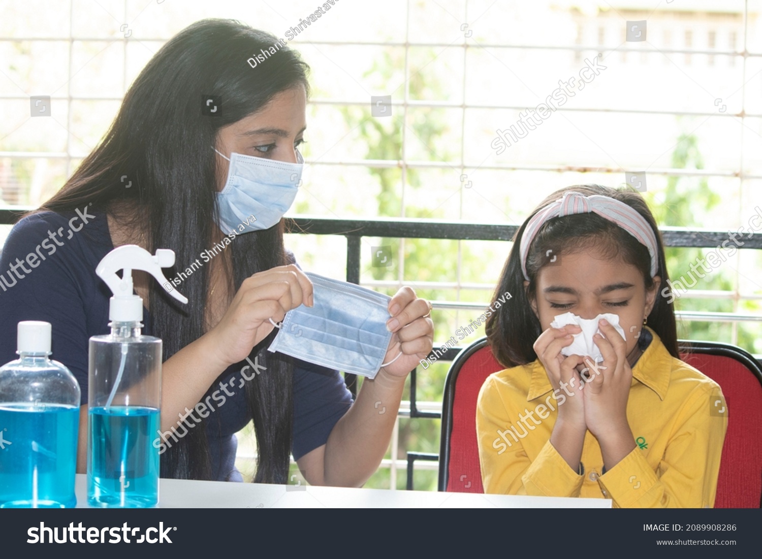 Cautious mother holding medical face mask for her daughter sneezing into tissue at home to protect self from infectious omicron variant of coronavirus disease #2089908286