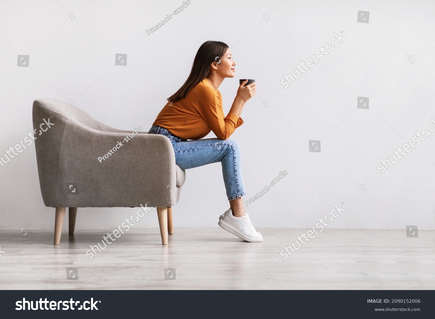 Side view of young Asian woman sitting in armchair, drinking hot aromatic coffee, relaxing against white studio wall, free space. Lovely millennial lady enjoying warm beverage on lazy day #2090152006