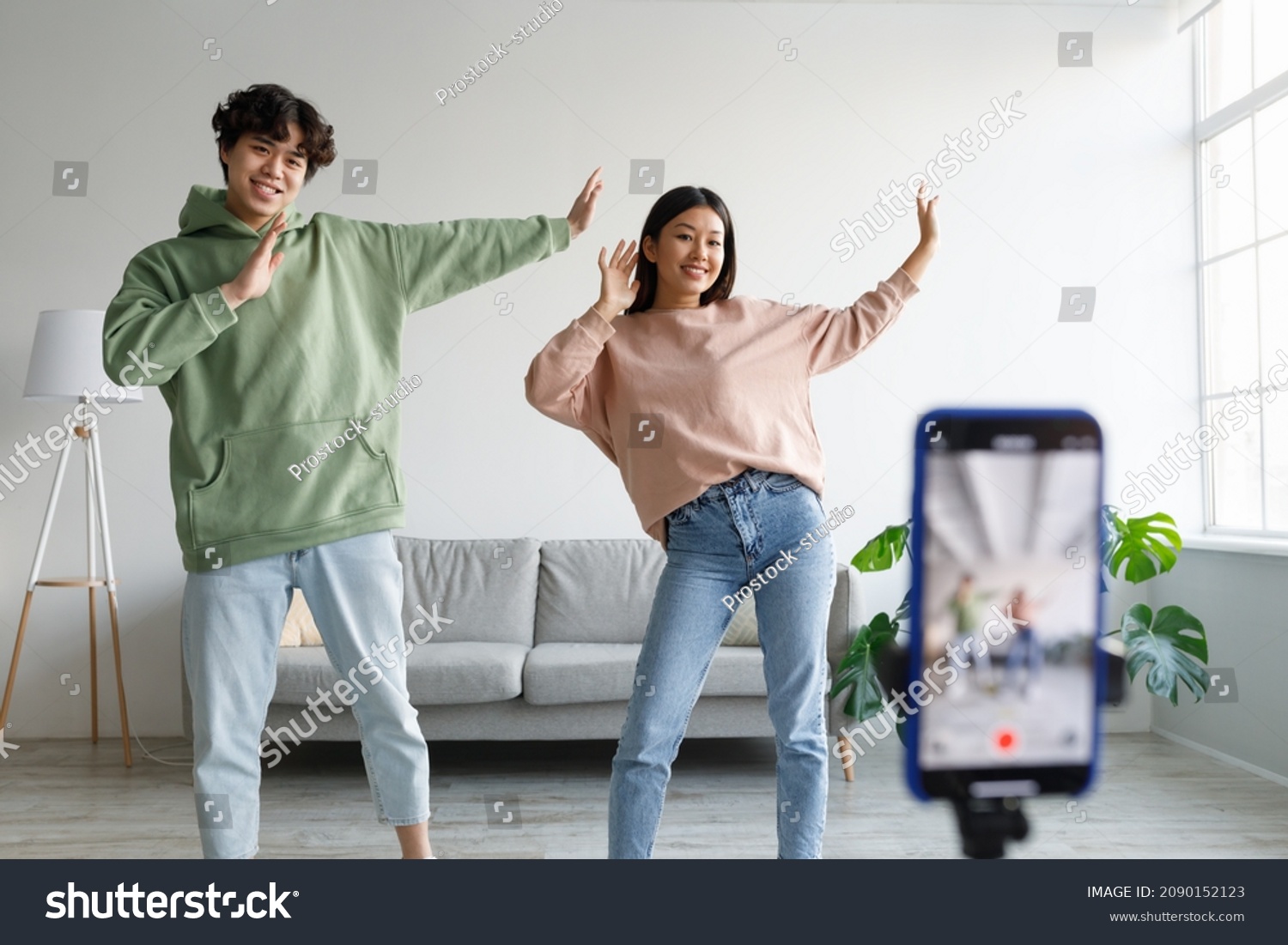 Young Asian couple recording video content, dancing on smartphone camera at home. Positive millennial boyfriend and girlfriend filming for social media, using mobile device. Vlogging concept #2090152123