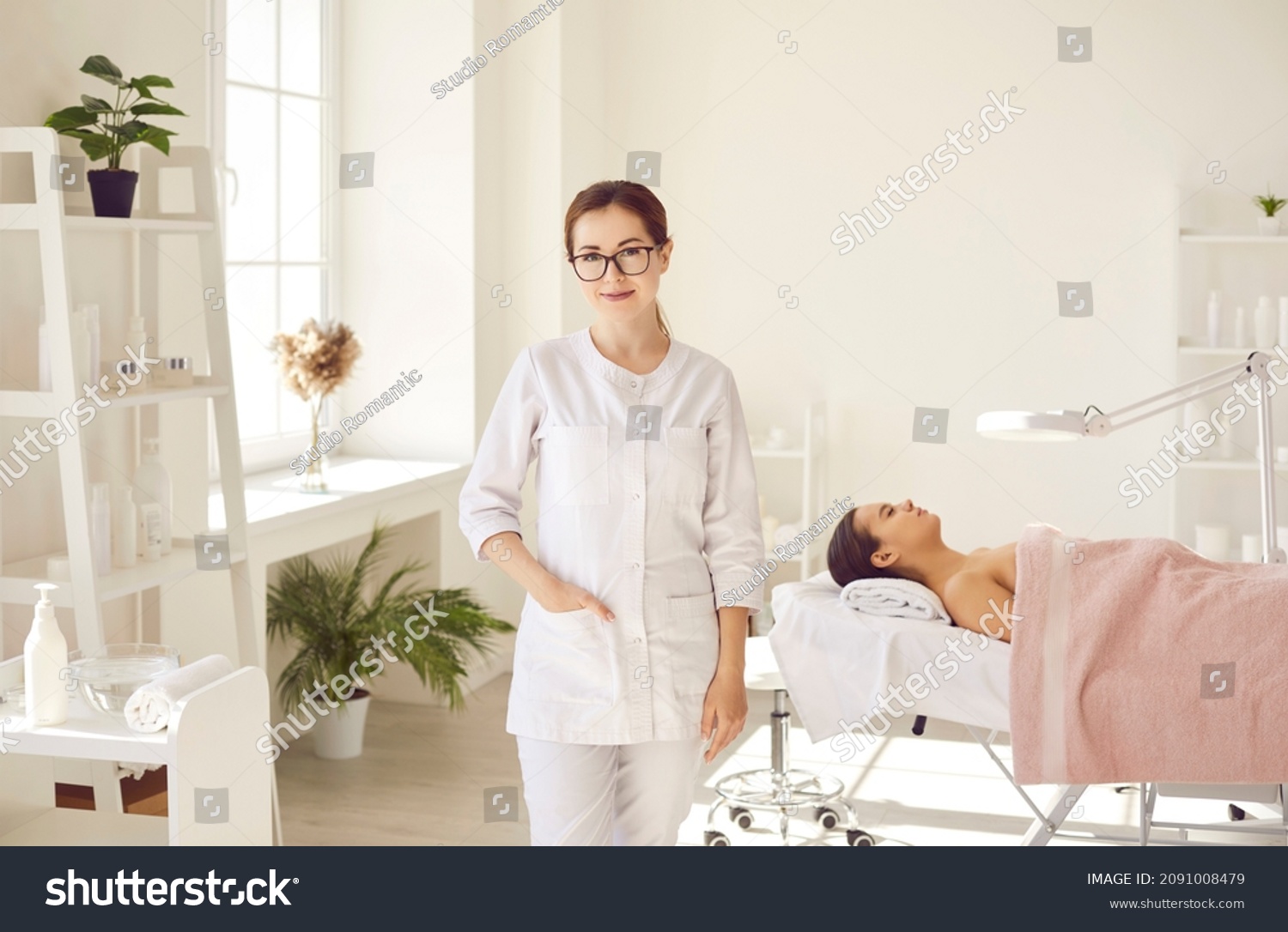 Female dermatologist, skin therapist, beautician and skincare professional with patient in office. Beautiful woman in coat uniform and glasses standing in room for examinations and cleaning procedures #2091008479