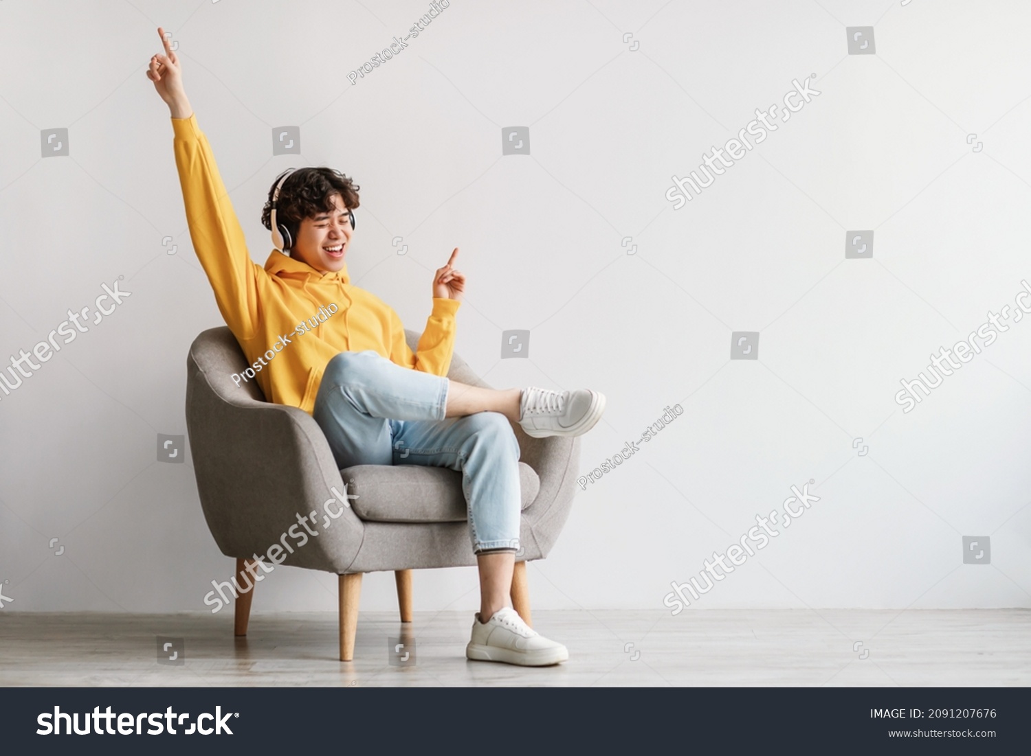 Joyful young Asian guy in wireless headphones sitting in armchair, having fun, listening to music, raising his arm, dancing to favorite song, enjoying cool soundtrack against white wall, empty space #2091207676