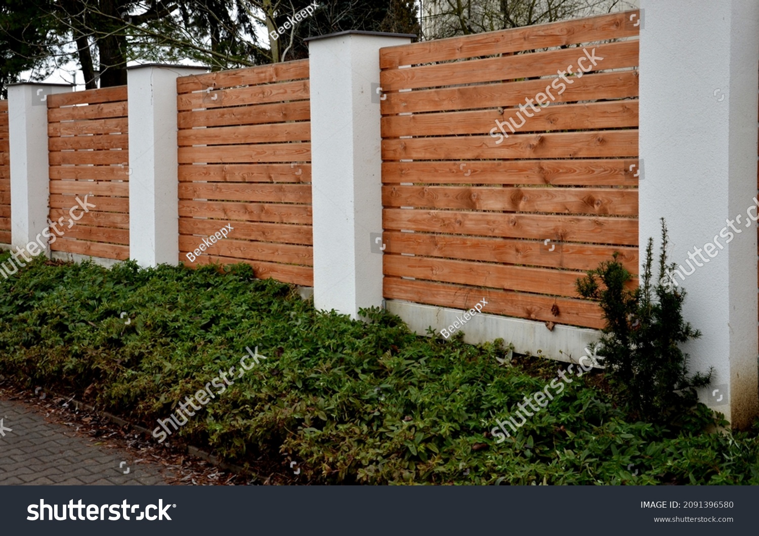 wooden fence made of natural planks. the columns are made of roughly plastered white columns. full fencing made of horizontally placed boards. adjacent to the land is a bed of shrubs #2091396580