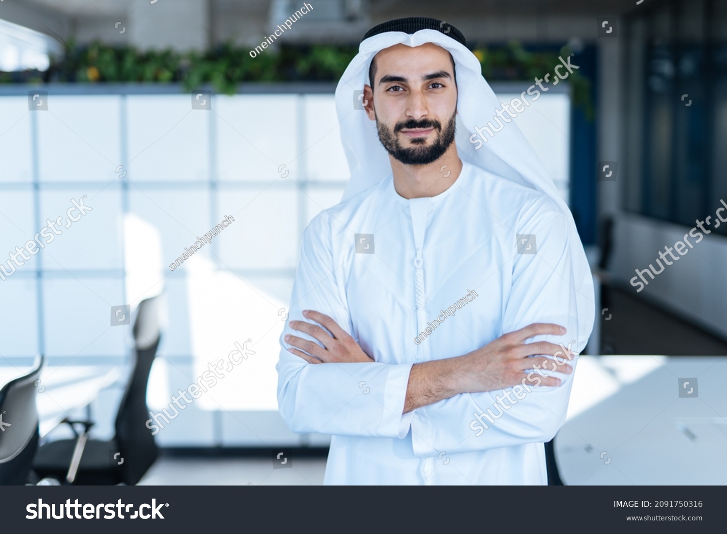 Handsome man with dish dasha working in his business office of Dubai. Portraits of a successful businessman in traditional emirates white dress. Concept about middle eastern cultures #2091750316