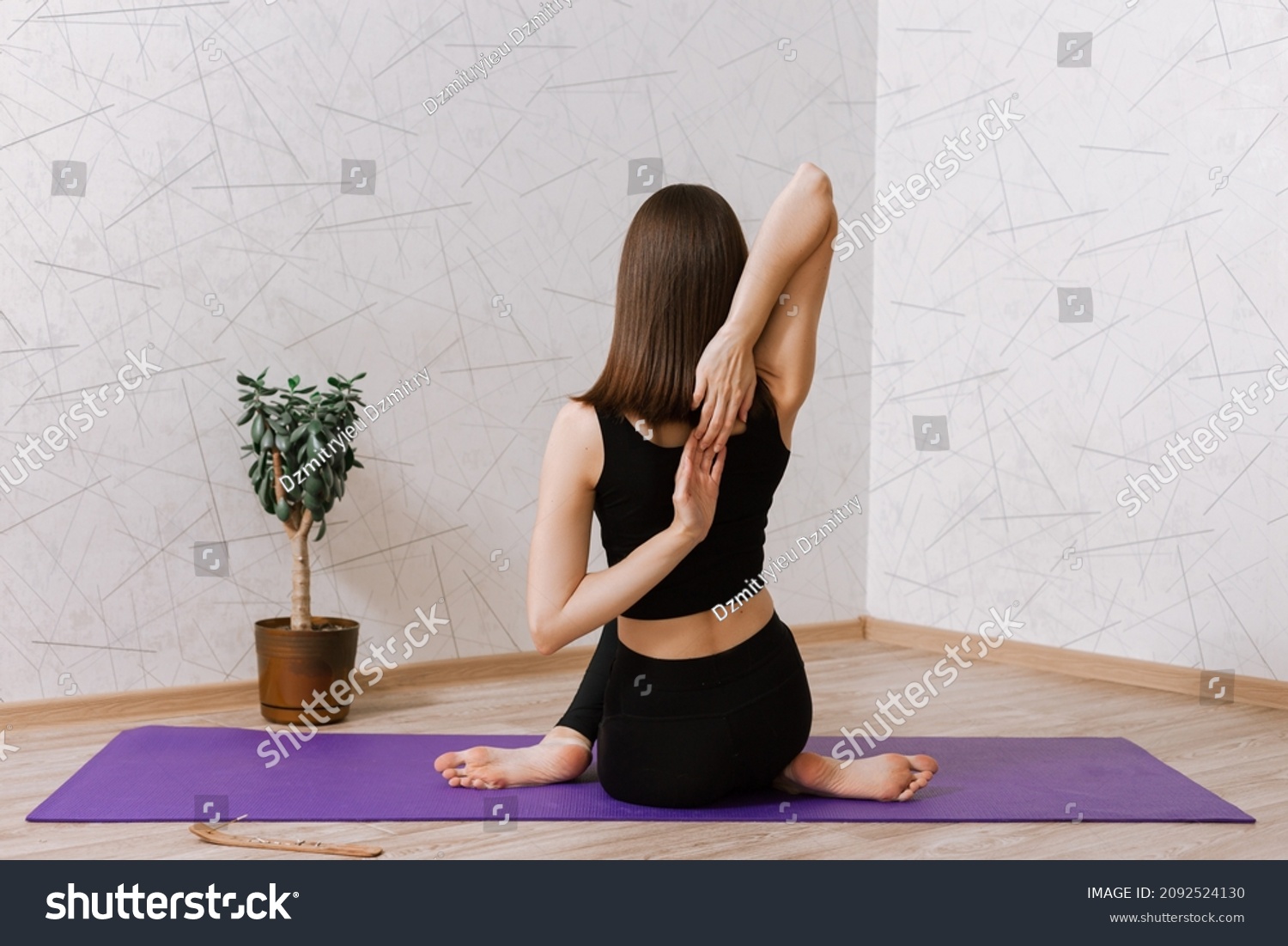 Back view of slim flexible woman practicing yoga in Cow Face Gomukhasana pose while sitting on mat in room at home  #2092524130