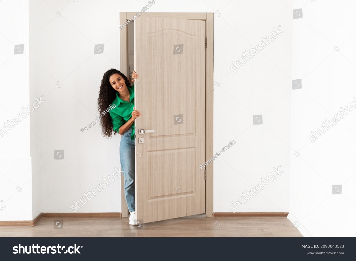 Portrait of cheerful young woman standing in doorway of modern apartment, millennial female homeowner holding slightly open ajar door looking out and smiling, greeting visitor, full body length #2093043523