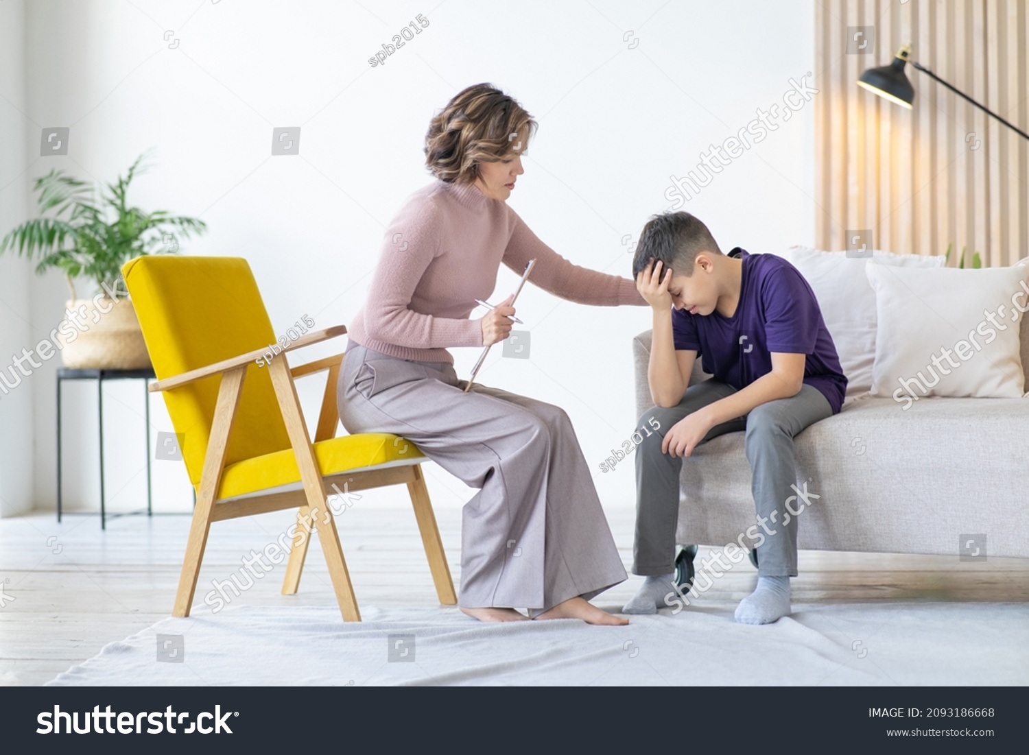 Counseling adolescent psychologist calms and holds adolescent by shoulder. Boy in purple t-shirt is worried and holds his head with his hand, while sitting on couch in minimalistic room. #2093186668