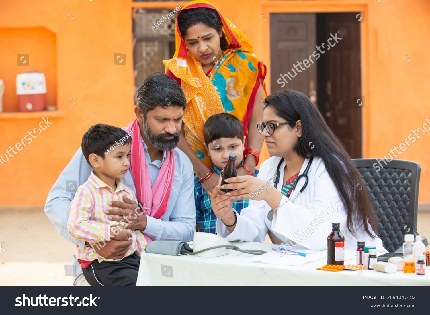 Young Indian Female Doctor with rural family give medicine outdoor village hospital,Healthcare government camp concept. Parents with two son getting medical help and support by pediatrician #2094047482