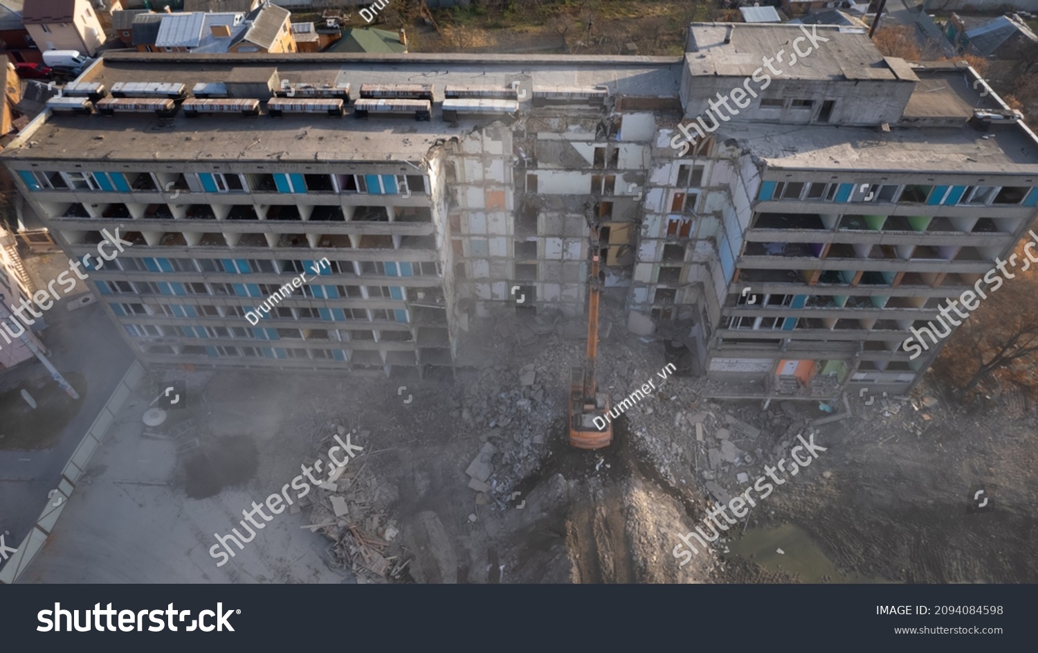 Excavator for dismantling buildings, using a special claw, dismantles a multi-storey building. The sun's rays break through a thick cloud of dust in front of the ruined building #2094084598
