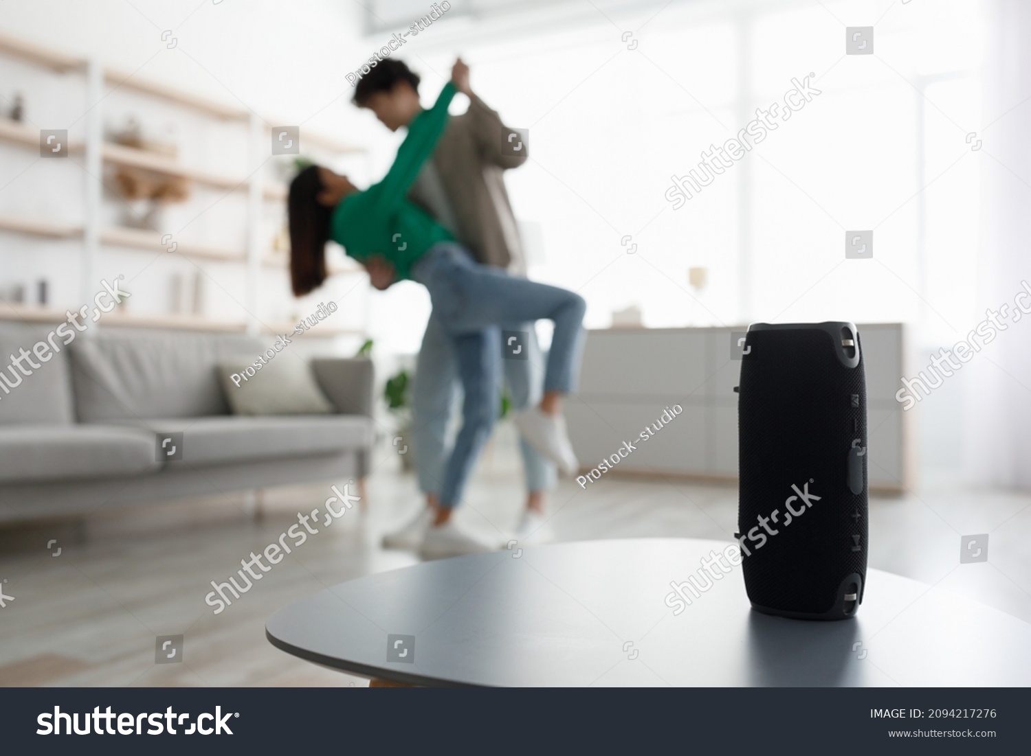 Millennial Asian couple dancing to popular music at home, selective focus on portable wireless speaker on table, copy space. Young family moving to their favorite song, using modern device #2094217276