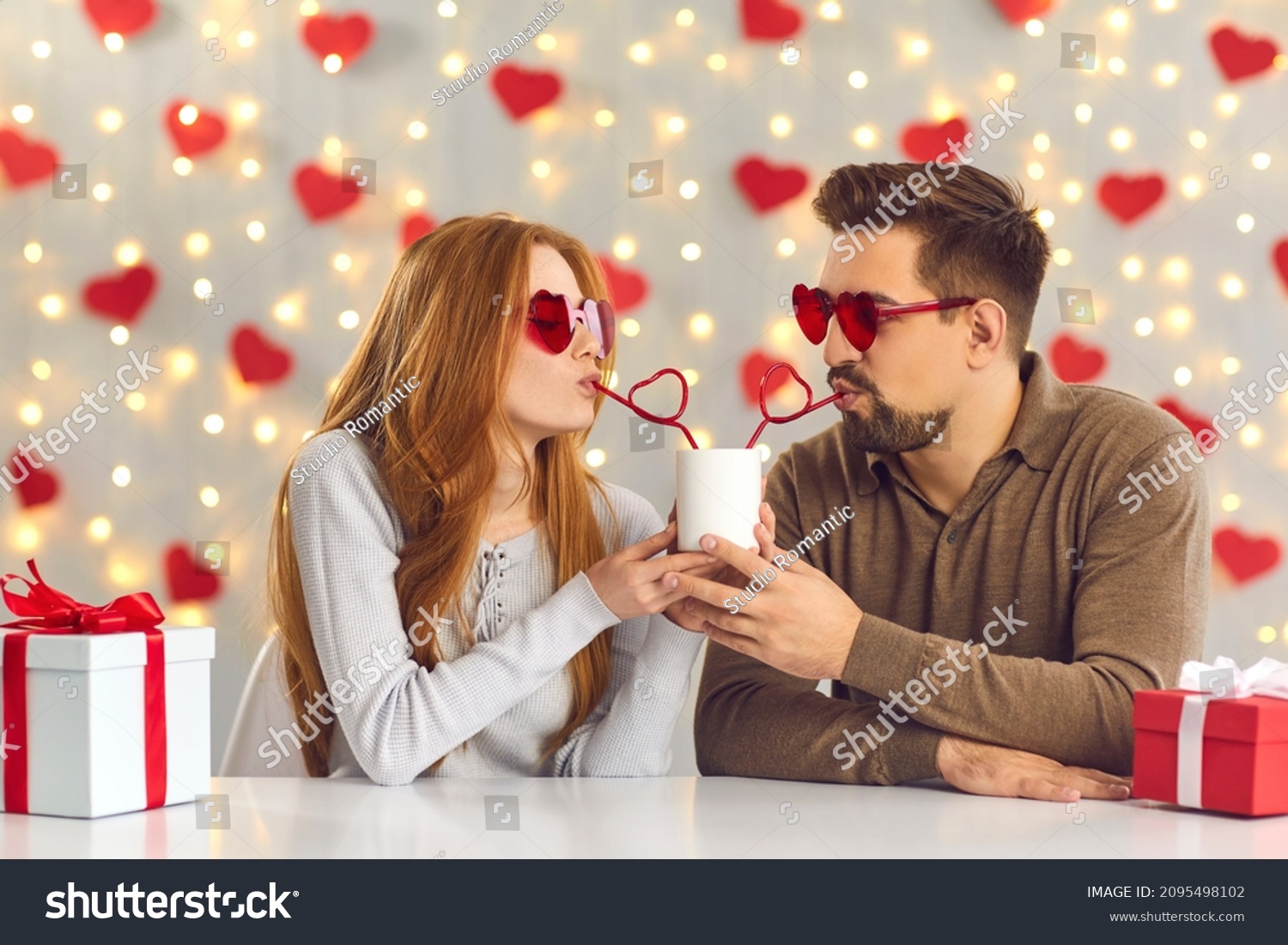 Young man and woman having a fun date on Saint Valentine's Day, sitting at table in cafe or at home, sipping drink from one cup through heart-shaped straws and enjoying cute and funny couple moment #2095498102