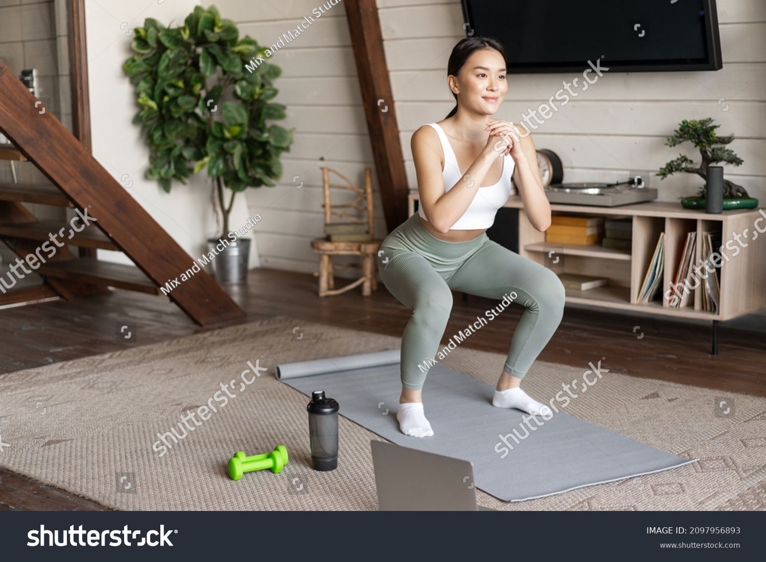 Young asian fitness girl doing squats workout at home, follow online sport gym instructor, standing on floor mat and smiling, exercising in living room #2097956893