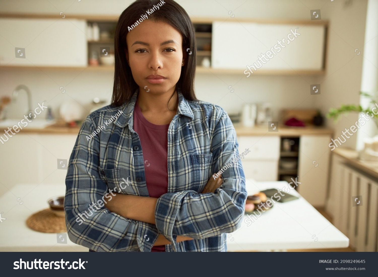 Suspicious mind. Beautiful middle-aged dark-skinned woman in blue plaid shirt standing with arms folded at kitchen looking at camera with mistrust, anger and grudge. Negative human emotions #2098249645