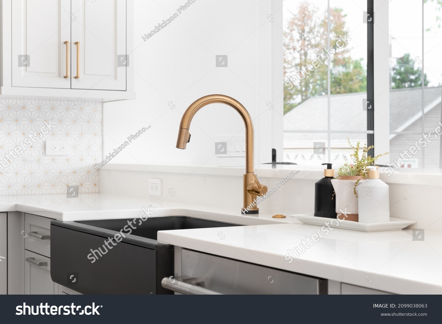 A beautiful sink in a remodeled modern farmhouse kitchen with a gold faucet, black farmhouse sink, white granite, and a tiled backsplash. No labels. #2099038063