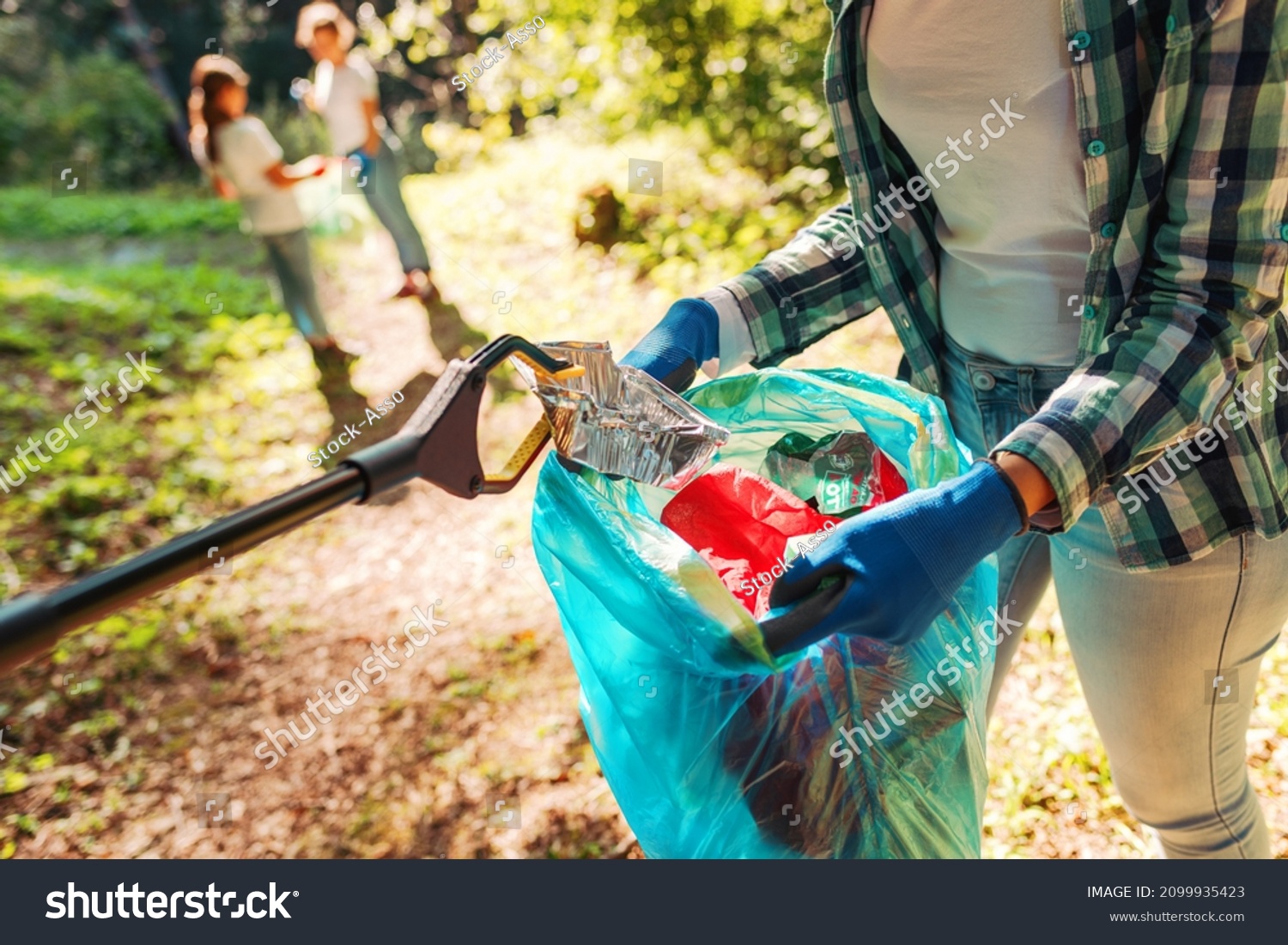 Volunteers cleaning up the park, a woman is putting trash in a garbage bag and some kids are helping her, environmental protection concept #2099935423