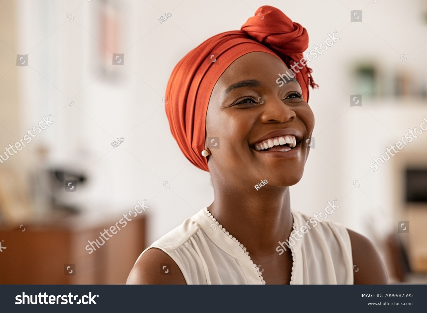 Smiling middle aged african american woman with orange headscarf. Beautiful black woman in casual clothing with traditional turban at home laughing. Portrait of mature carefree lady looking away. #2099982595
