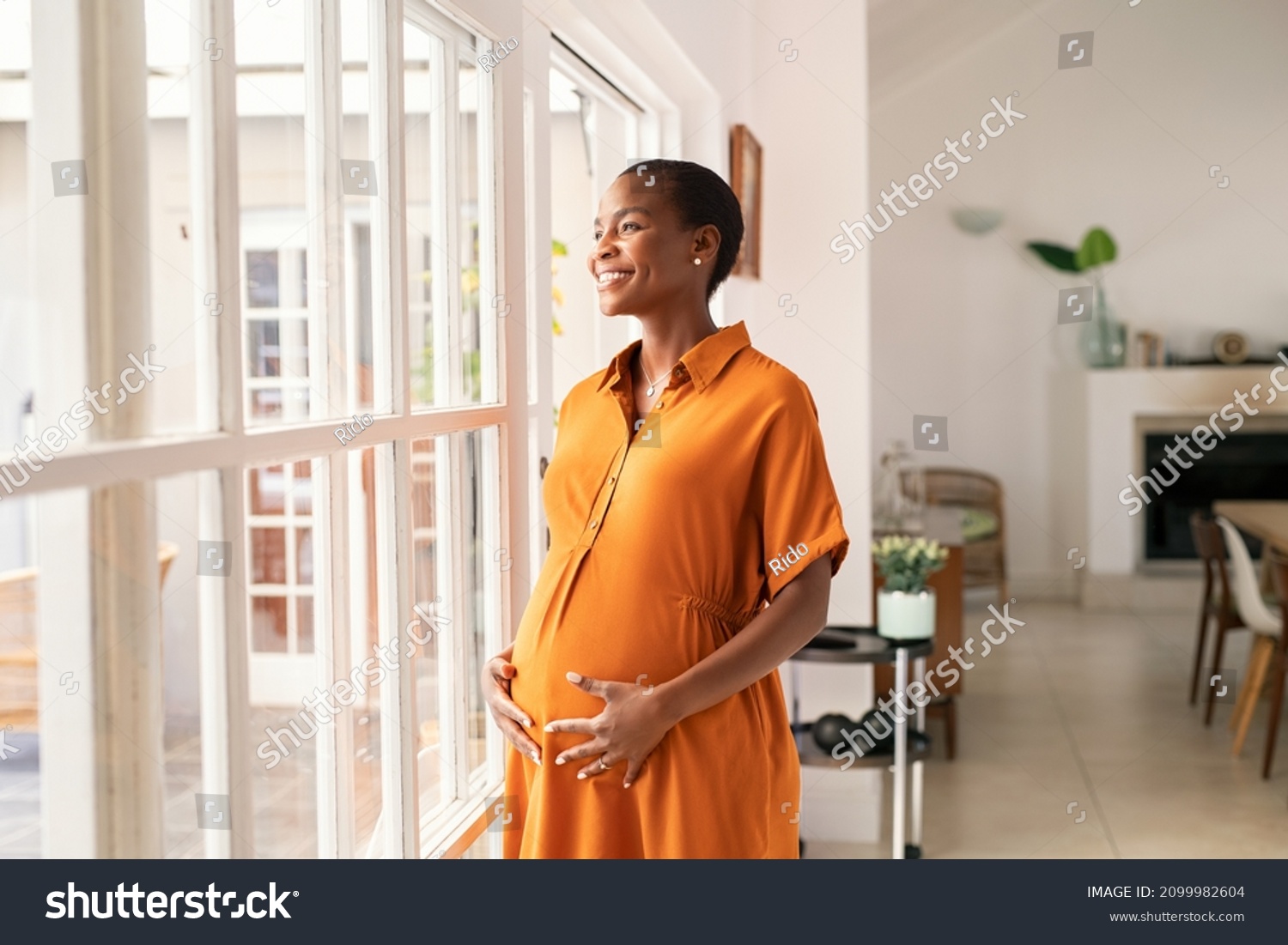 Mid black pregnant woman standing near window at home and thinking about her future family. Smiling african american lady with hands on belly imagine the growth of his baby. Middle aged pregnant woman #2099982604