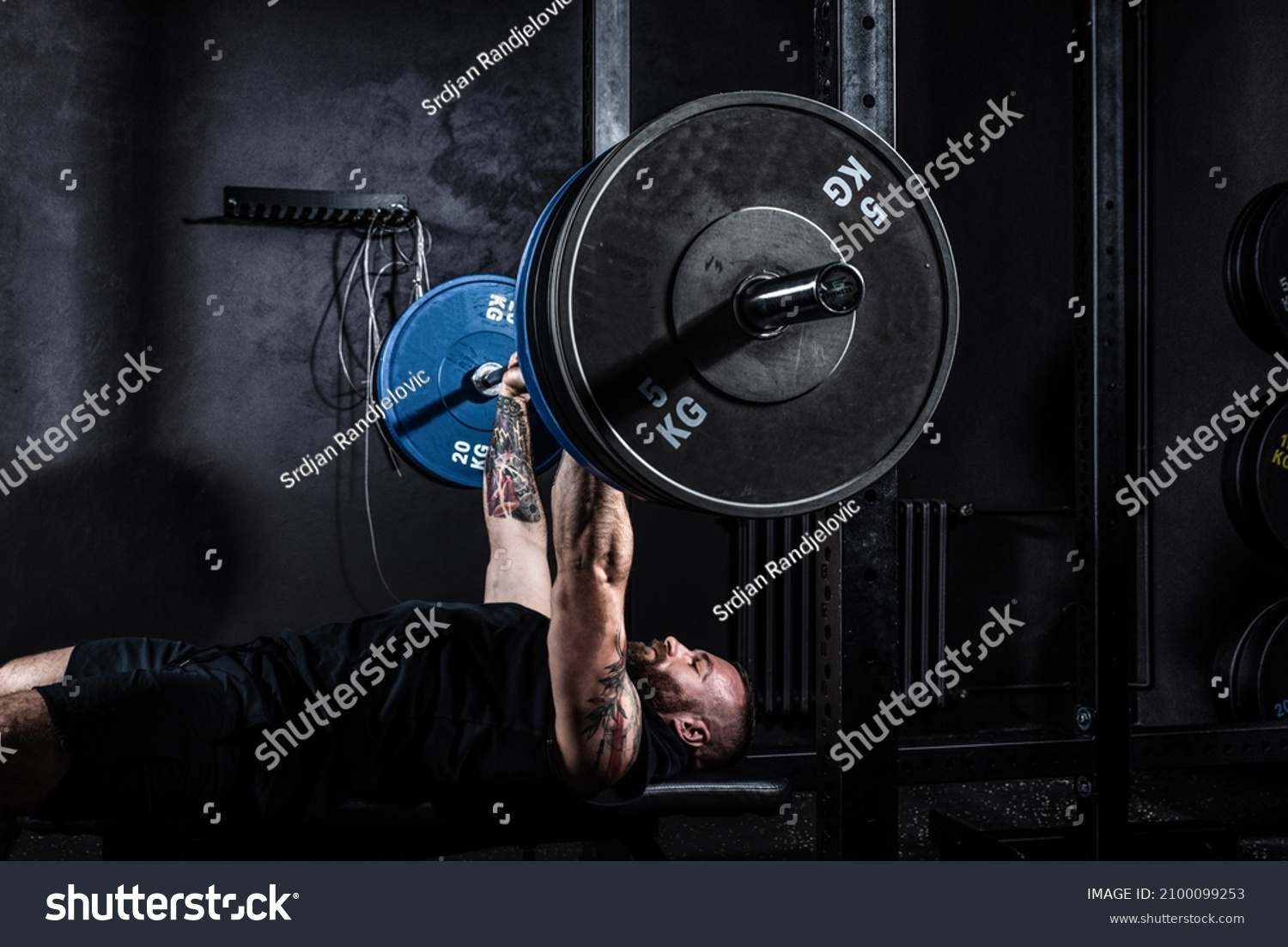 Strong sweaty muscular athlete fit active man doing hardcore bench press with heavy barbell weights in the gym. Active male bodybuilder workout and crossfit training concept. Real people exercise #2100099253