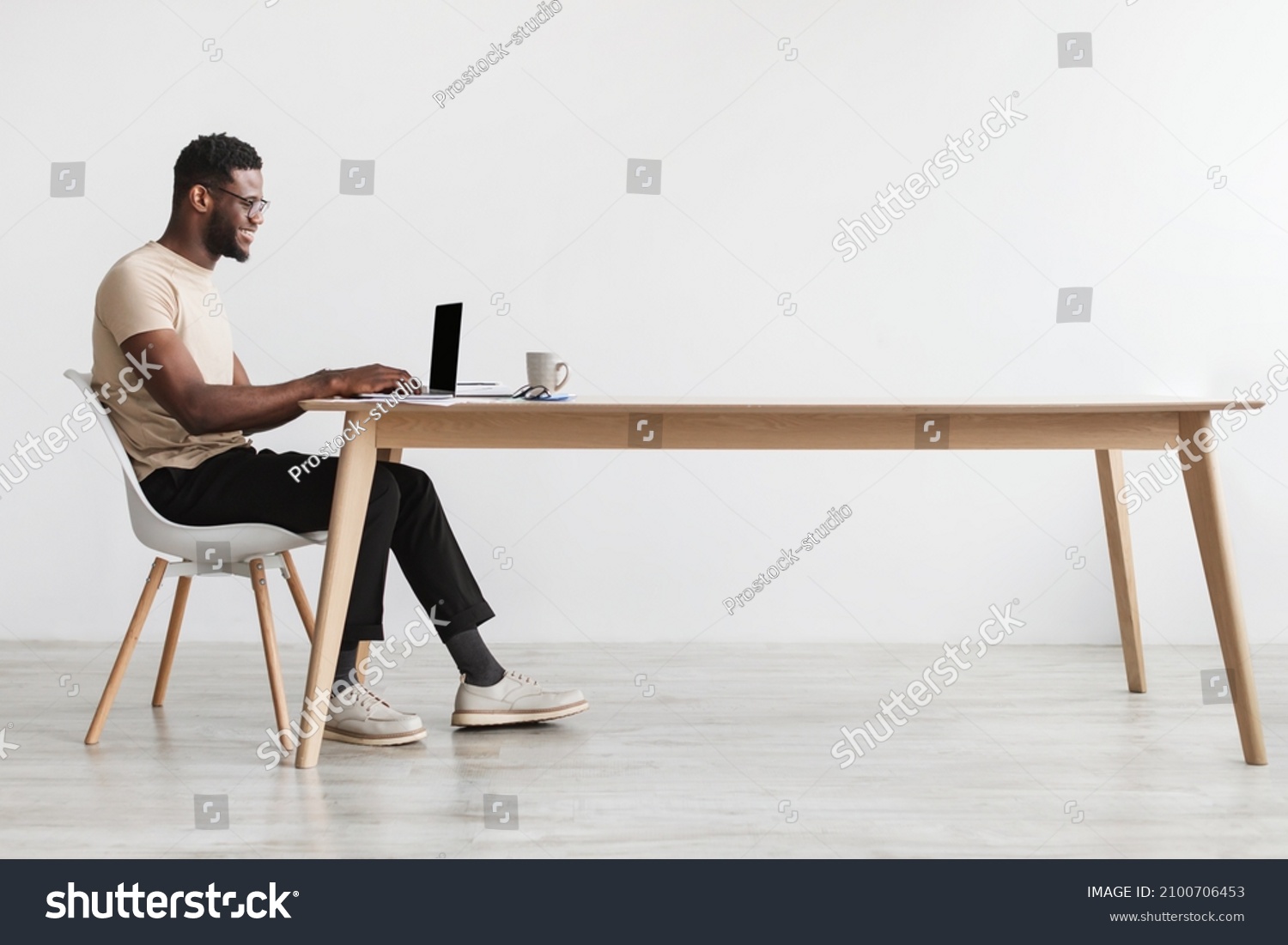 Side view of young black guy in casual wear using laptop, sitting at desk, working online from home office, copy space. Millennial African American man freelancing, having remote job #2100706453