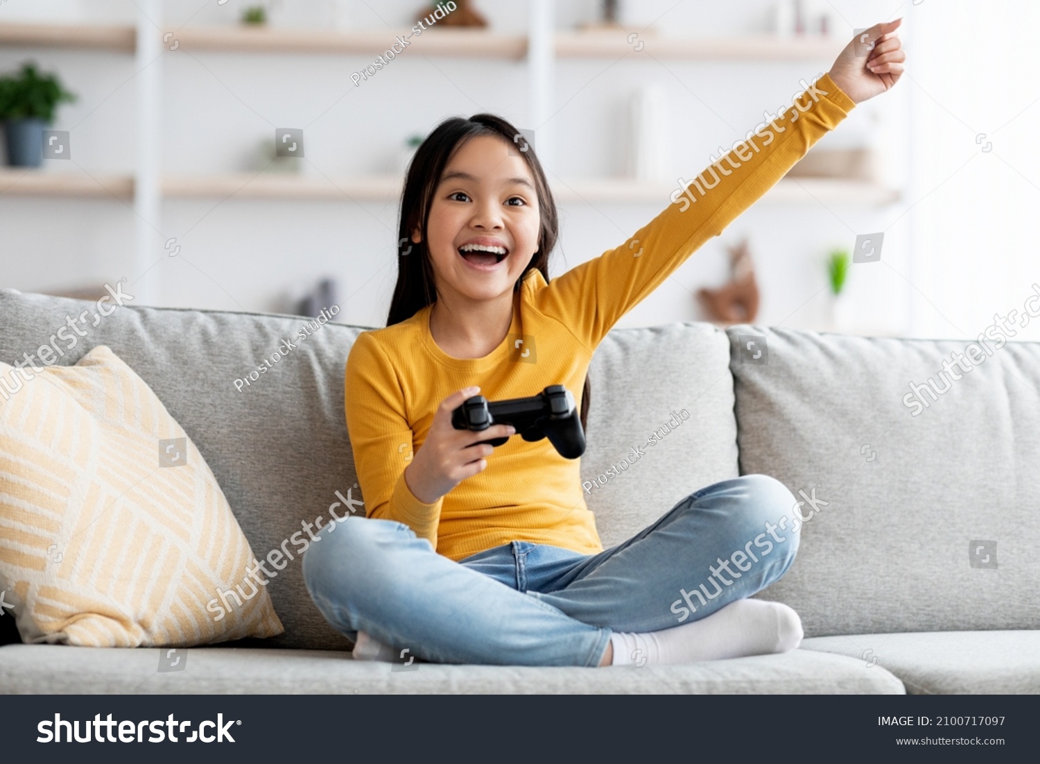 Happy cute asian girl schooler sitting on couch in living room, holding joystick, playing handheld video game and gesturing, enjoying weekend at home, kids entertaining concept, copy space #2100717097