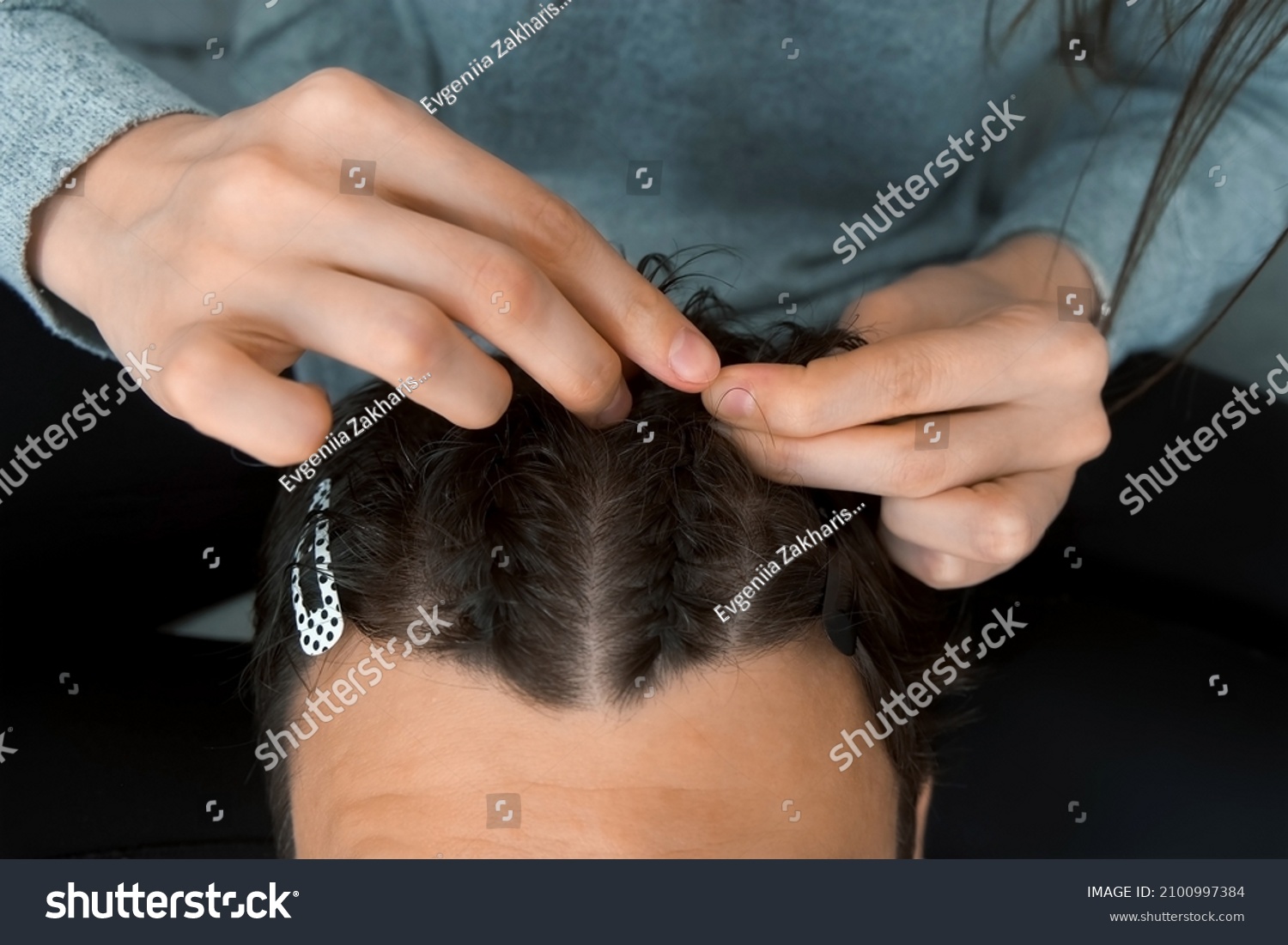 Woman is doing french braid on short men's hair, closeup view. She is doing a man's hair while sitting at home on the couch. Evening of a young married couple together. #2100997384