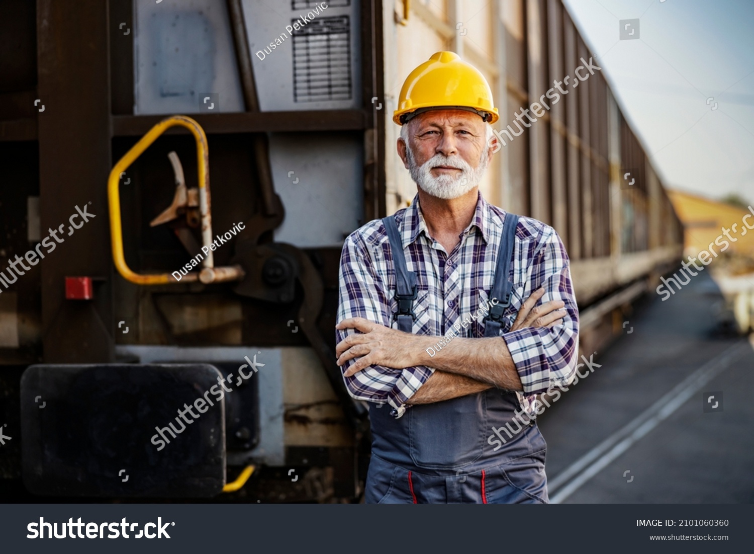 Industry transportation. A senior worker standing next to a wagon. A proud senior worker with a protective helmet on his head is standing with arms crossed next to a wagon. #2101060360