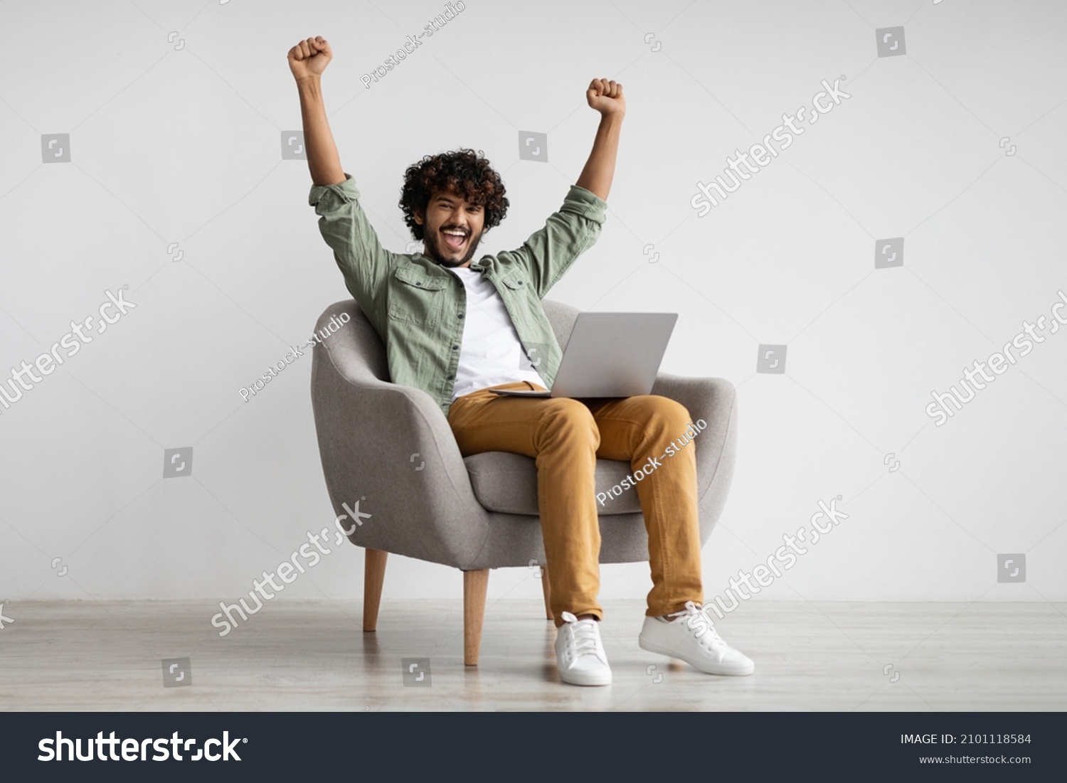 Emotional curly young hindu guy in casual sitting in armchair with modern laptop and raising hands up, celebrating success, trading on stocks and markets online, got good profit, copy space #2101118584