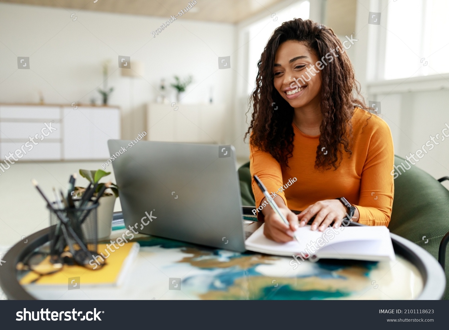 Smiling young African American woman sitting at desk working on laptop taking notes in notebook, happy millennial female studying online, watching webinar using computer and writing check list #2101118623
