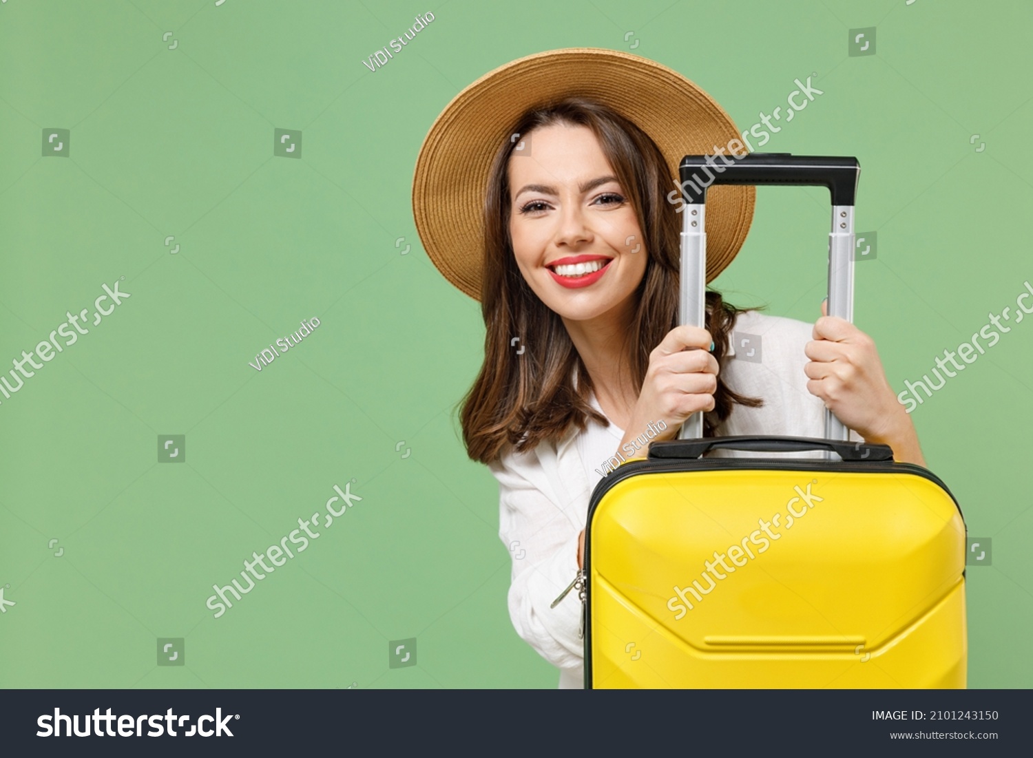 Close up cheerful fun traveler tourist woman in casual clothes hat hiding with yellow suitcase valise isolated on green background Passenger travel abroad weekends getaway Air flight journey concept. #2101243150