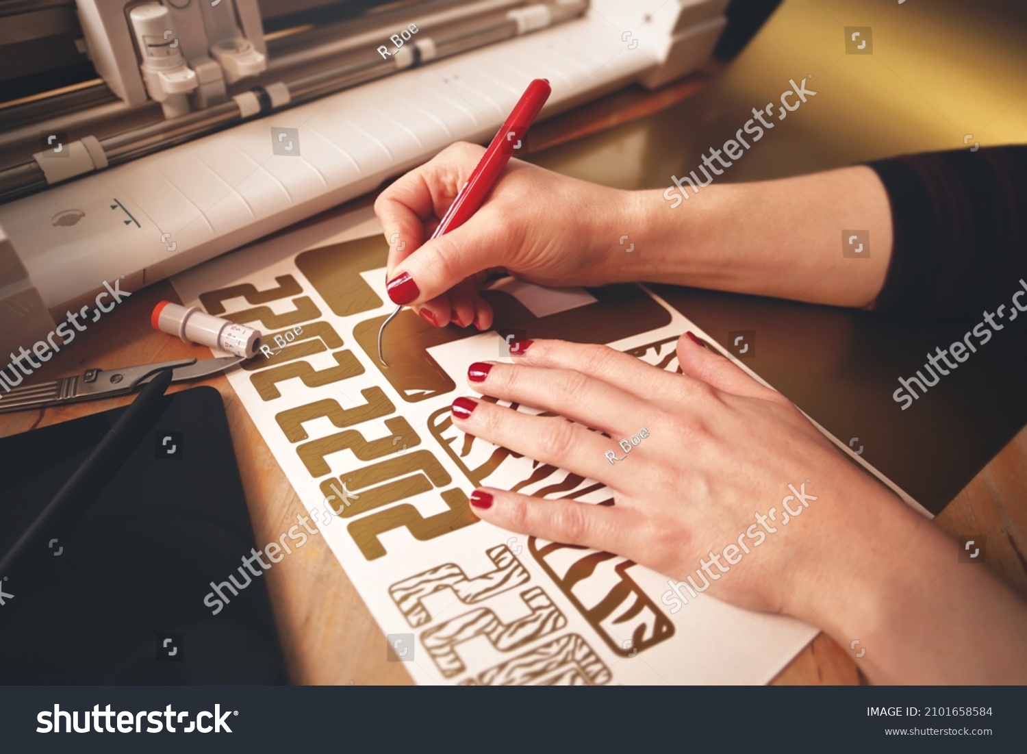woman with red painted nails removes excess material with weeding tool to make 2022 tiger design stickers from gold vinyl foil. workplace with cutting machine, tools, tablet computer. selective focus #2101658584