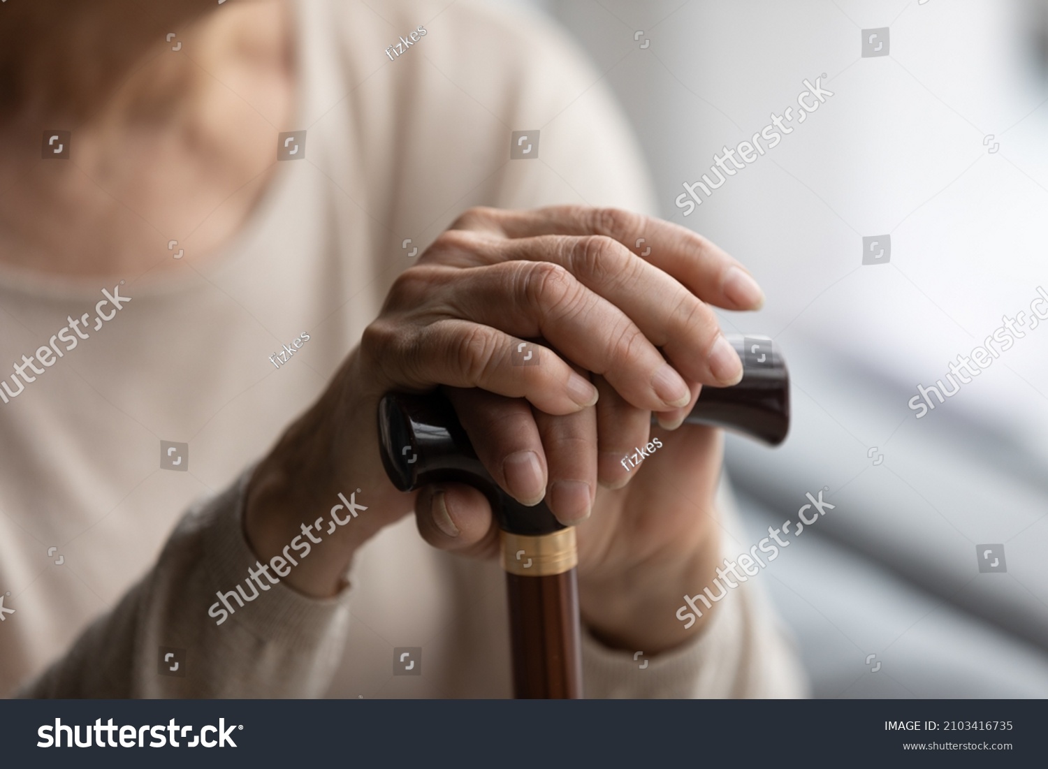 Close up focus on folded wrinkled female hands on wooden cane. Cropped thoughtful elderly senior grandmother having walking disability, using stick indoors, old retired people lifestyle concept. #2103416735