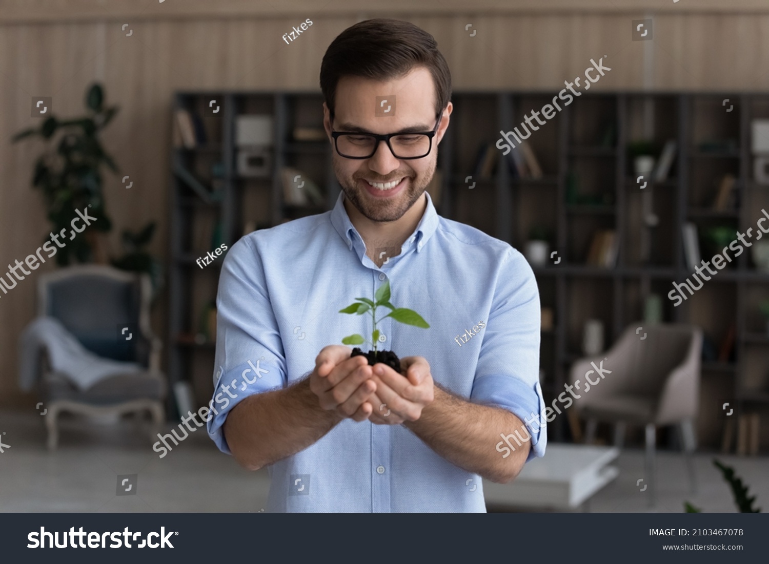 Smiling businessman wearing glasses holding small green plant sprout with soil, standing, happy entrepreneur employee with growing tree, startup project, profit, investment and growth concept #2103467078