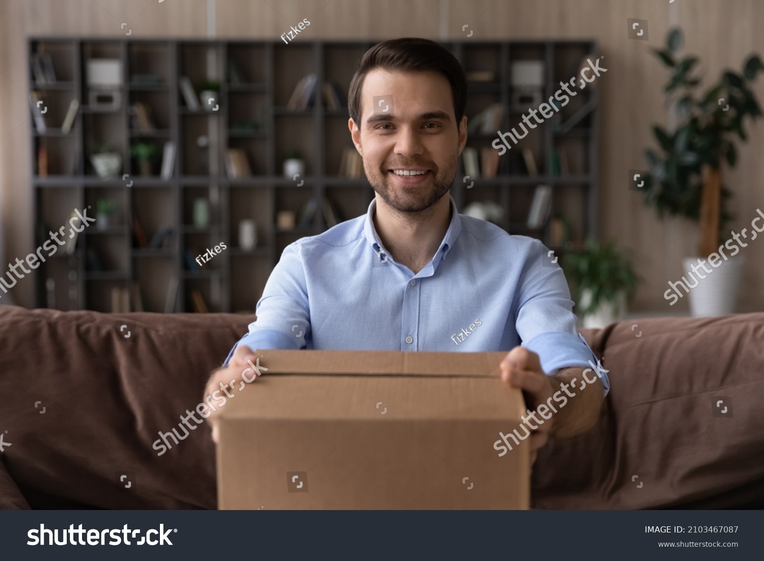 Head shot portrait smiling man holding cardboard box, giving or receiving parcel, sitting on couch at home, happy satisfied customer looking at camera, good delivery service and fast shipping concept #2103467087