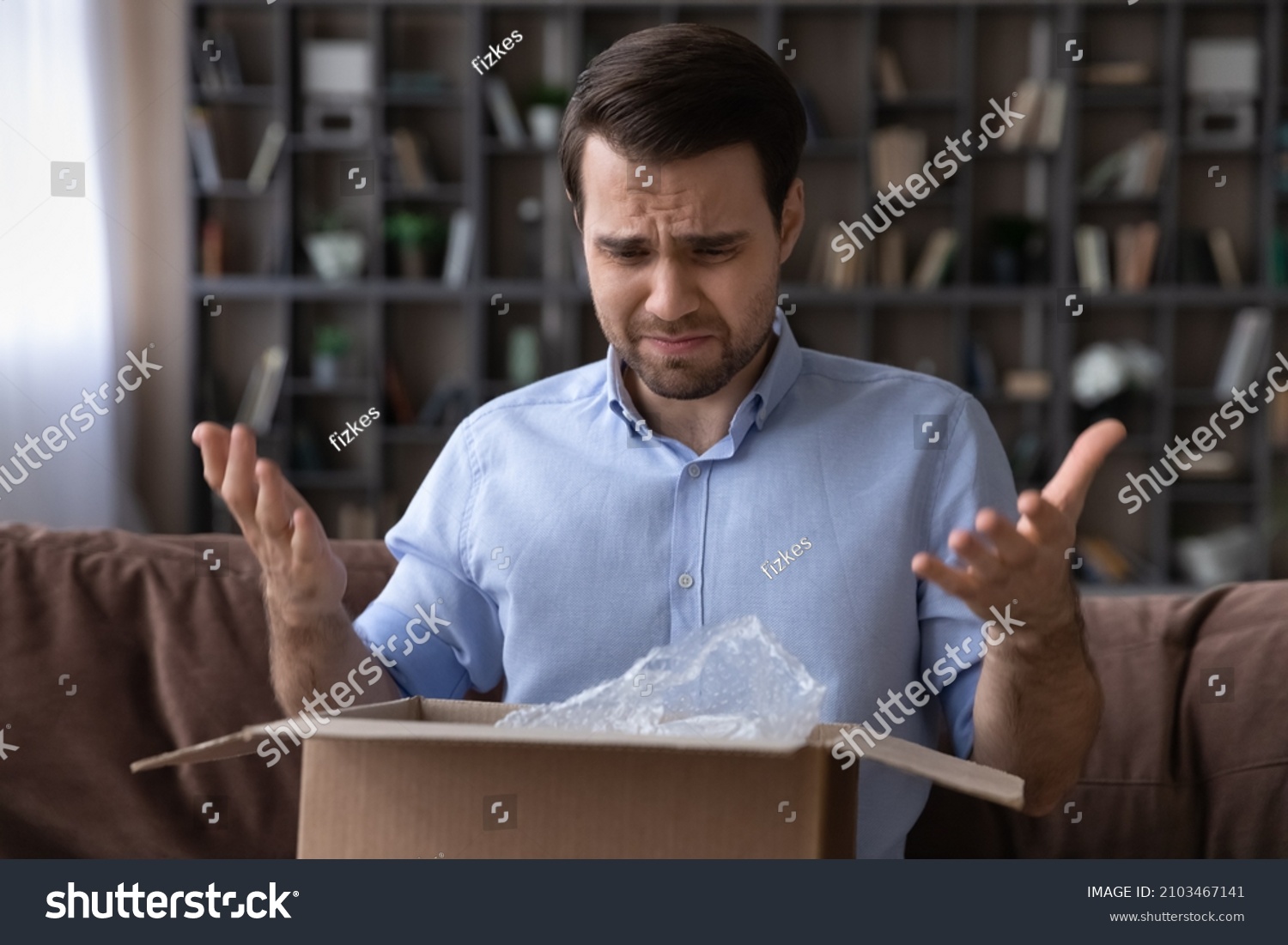 Head shot unhappy dissatisfied man opening parcel at home, sitting on couch with cardboard box, angry displeased customer confused by wrong or damaged order, bad delivery shipping service concept #2103467141