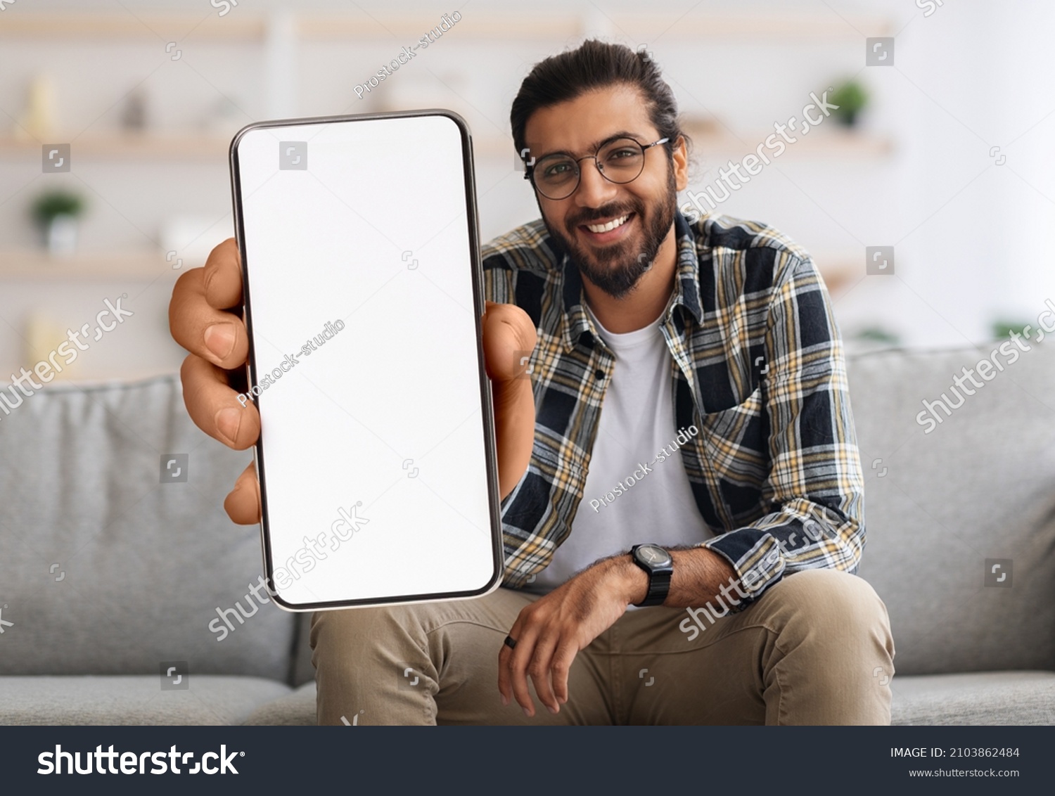 Cheerful Indian Guy Showing Smartphone With Big Blank White Screen At Camera, Happy Young Eastern Man Recommending New Mobile App Or Website While Sitting On Couch At Home, Creative Collage, Mockup #2103862484