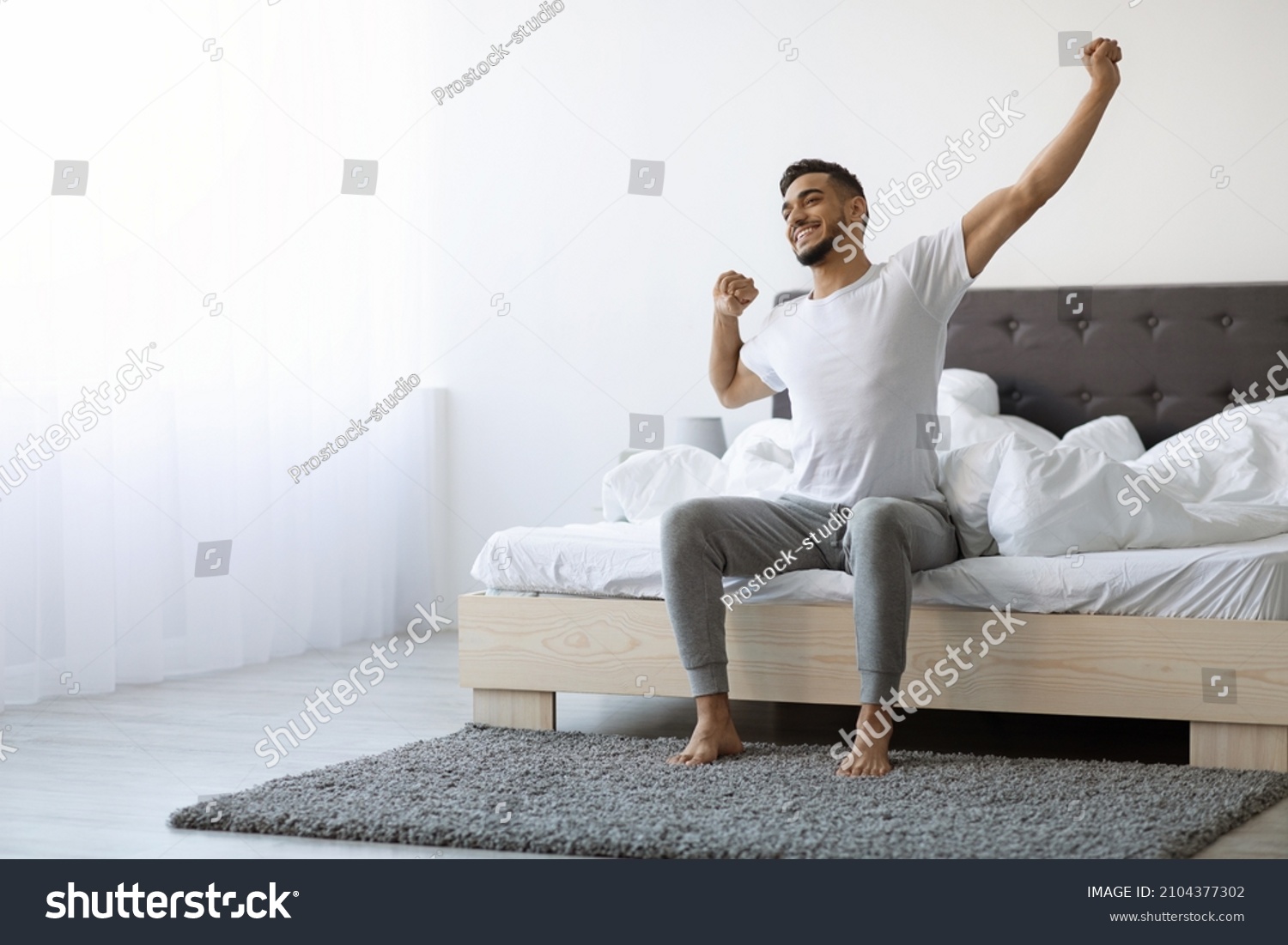 Good Morning. Happy Millennial Arab Guy Sitting On Bed And Stretching After Good Sleep, Handsome Smiling Young Middle Eastern Man Relaxing In Cozy Bedroom, Enjoying Home Comfort, Copy Space #2104377302