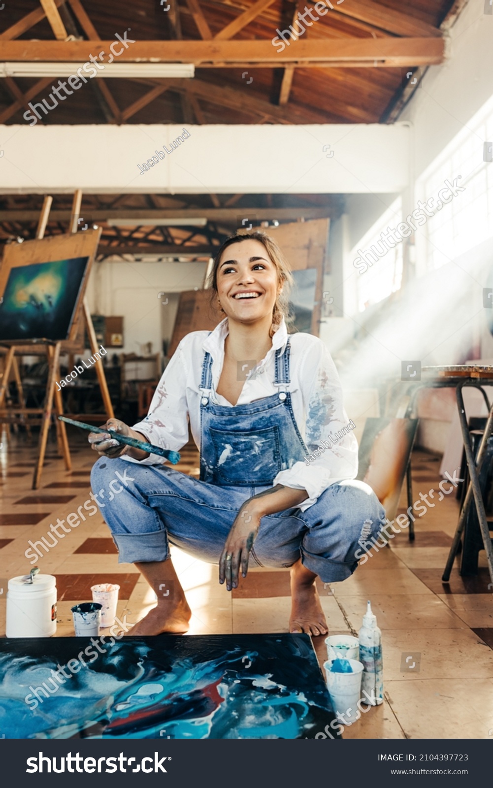 Inspired young artist looking away with a smile in her art studio. Happy female painter contemplating new creative ideas for her art project. Imaginative young woman painting on a canvas on the floor. #2104397723