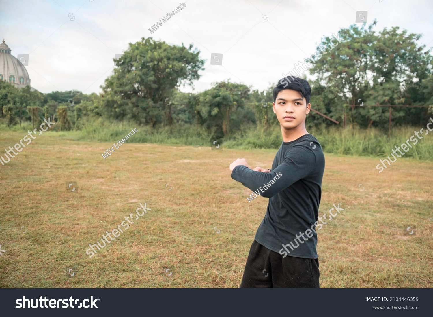 A young asian man doing standing oblique twists. In preparation for a morning run at an outdoor field. #2104446359