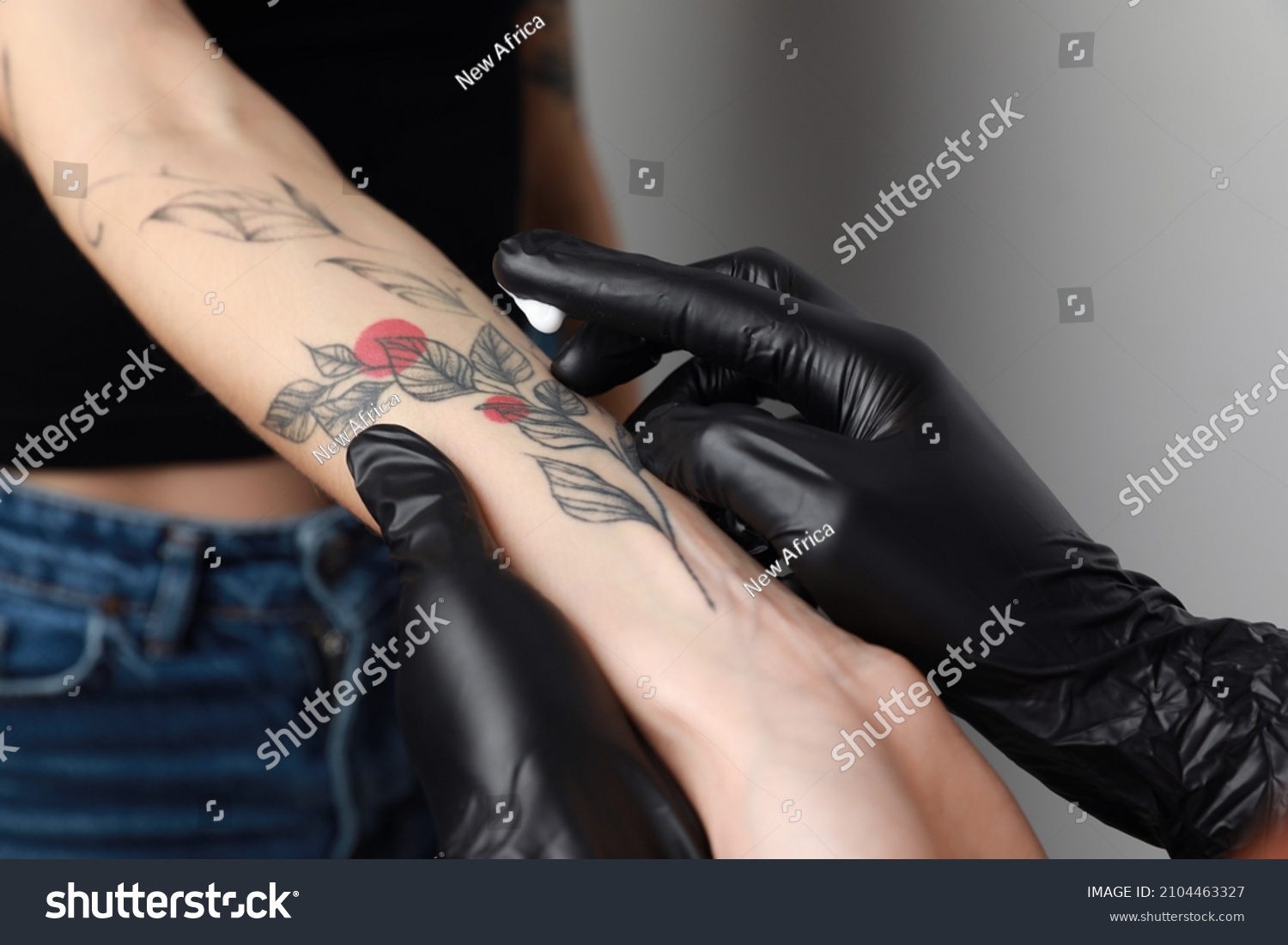 Worker in gloves applying cream on woman's arm with tattoo against light background, closeup #2104463327