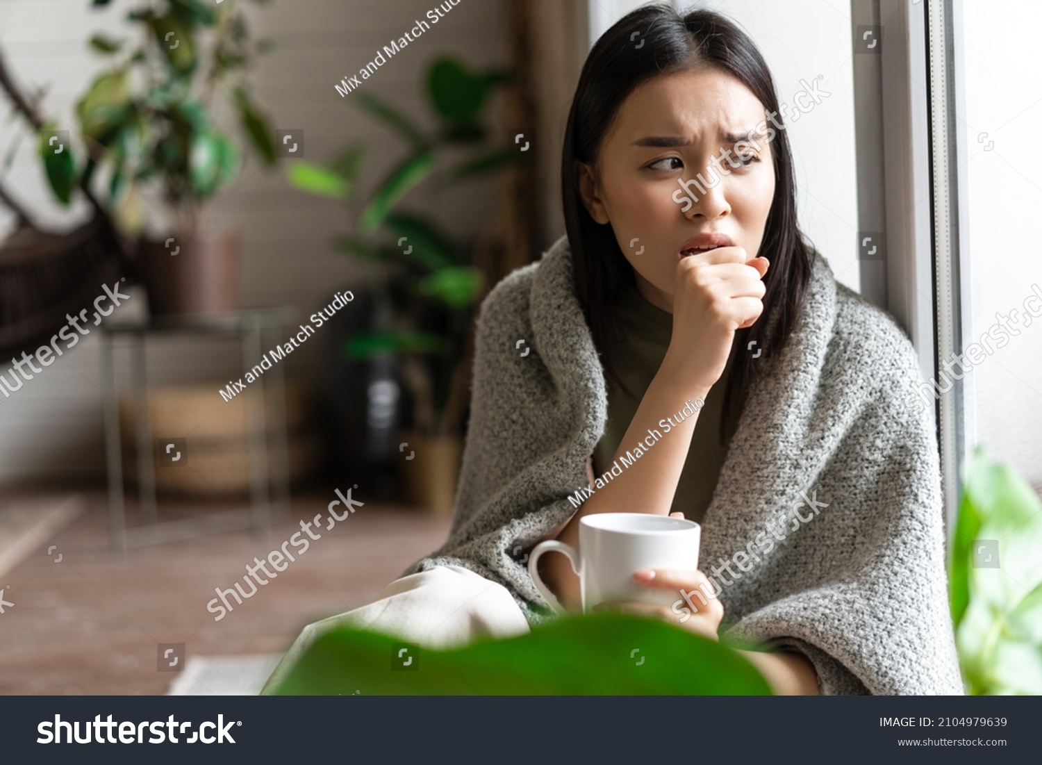 Sad ill asian girl staying on self quarantine during covid, catching flu and sitting at home with cup of tea near window, coughing in hand #2104979639