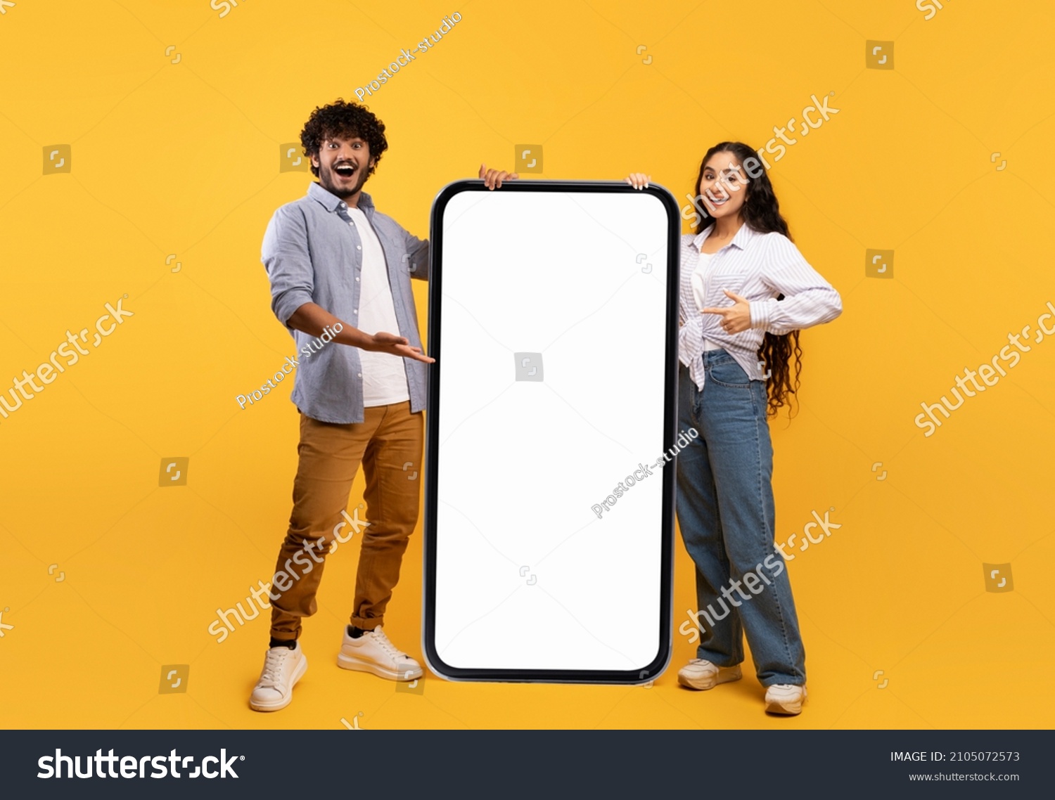 Cool mobile offer. Happy indian couple pointing at big cellphone with white screen for mockup, demonstrating free space for phone application or website design, yellow background #2105072573