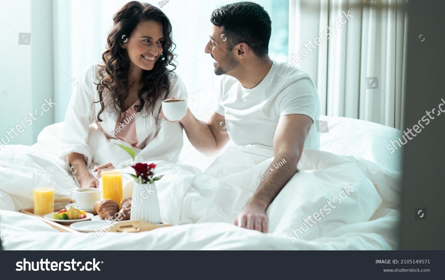 Isolated Beautiful Ethnic Couple Celebrating Valentines Day in a Fancy Hotel Suite, Eating Breakfast Off of a Fancy Tray As They Stay Under The Blanket, Looking Into Each Others Eyes and Smiling. #2105149571
