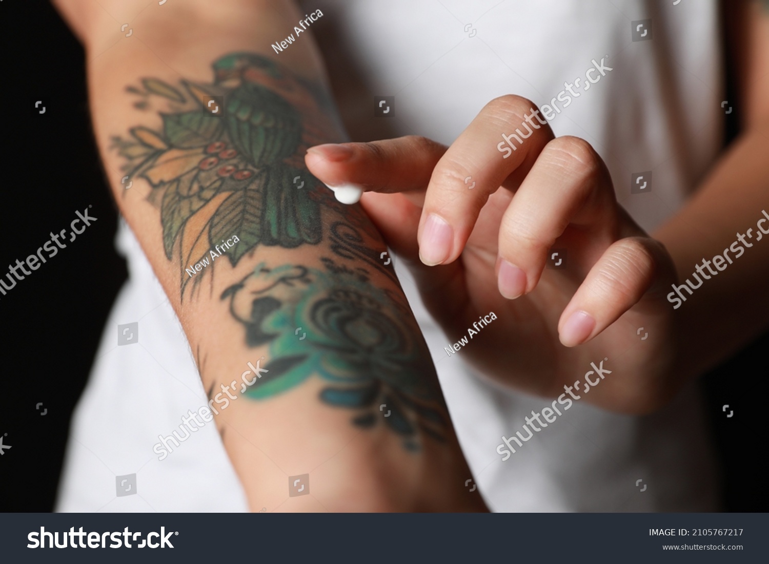 Woman applying cream on her arm with tattoos against black background, closeup #2105767217