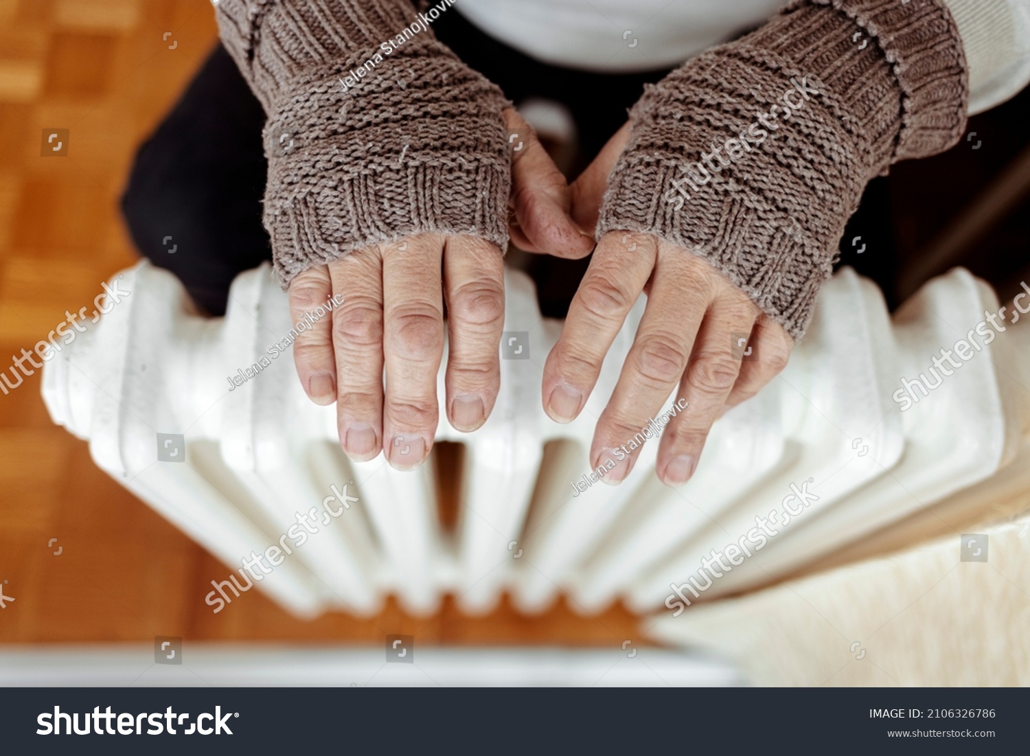 A man hands in wool gloves warm near the heater. Old men's hands in knitted gloves on heating radiator at home during the day. Person heating their hands at home over a domestic radiator in winter. #2106326786