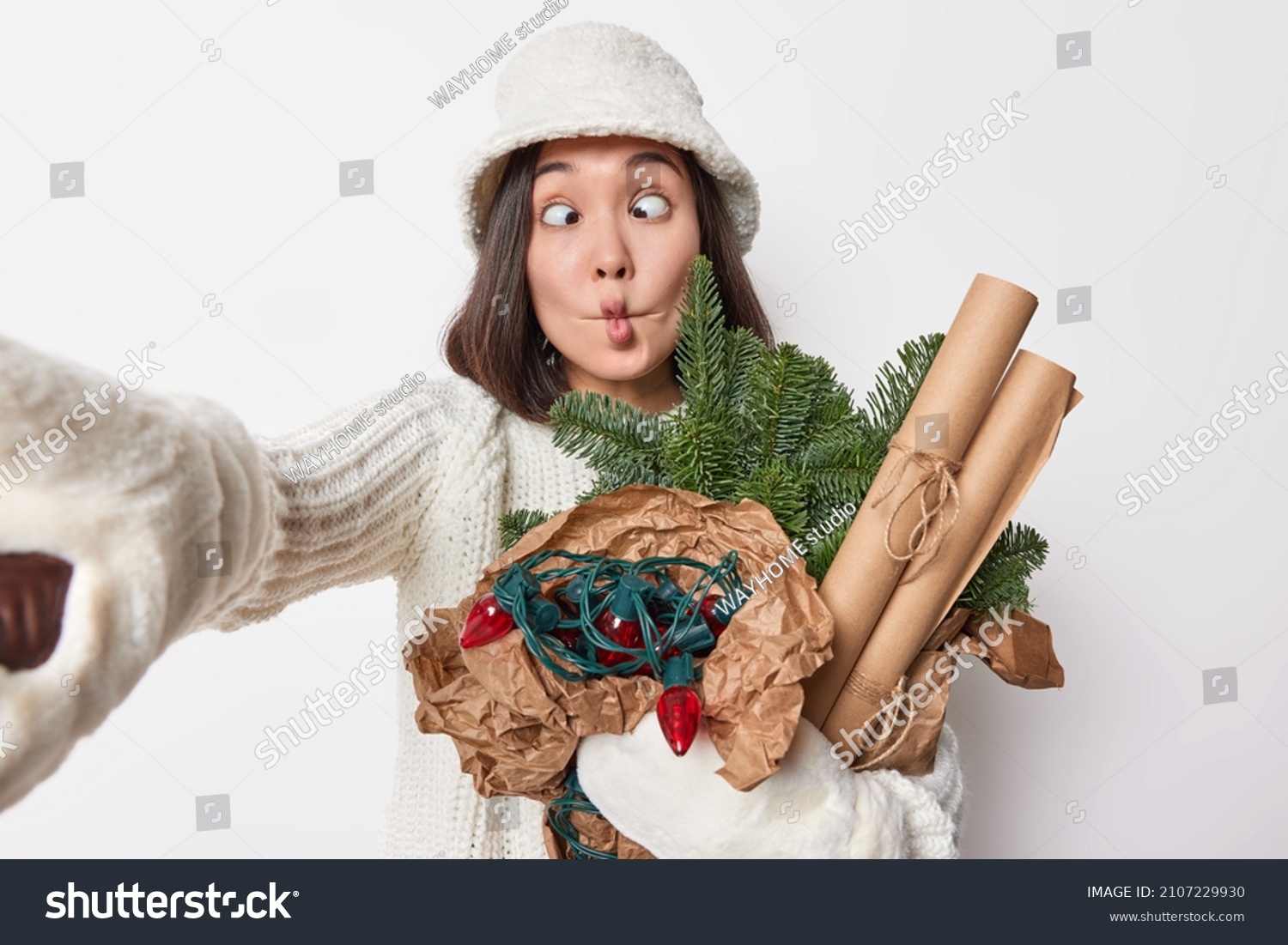 Funny brunette young woman makes fish lips crosses eyes foolishes around shows grimace keeps arm outstretched for making selfie wears white panama warm knitted sweater prepares for New Year. #2107229930