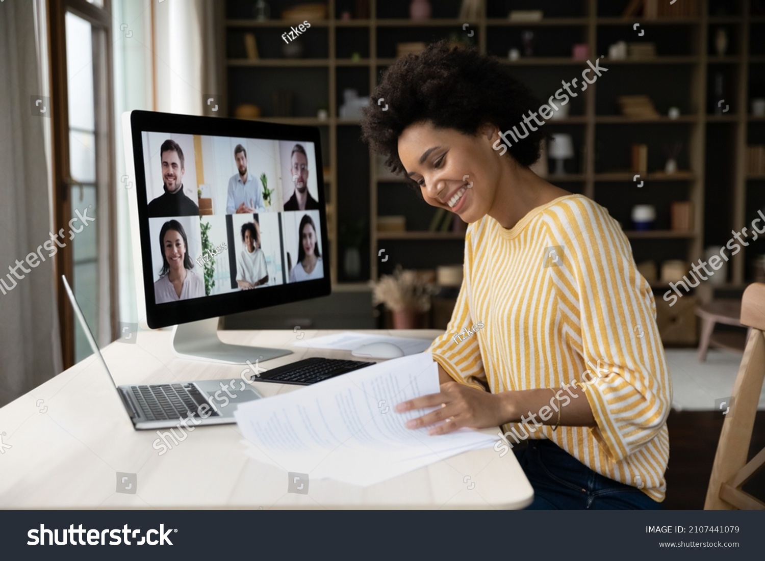 Side view focused smiling young African American female employee looking through paper documents, holding video conference call negotiations meeting with diverse colleagues business people online. #2107441079