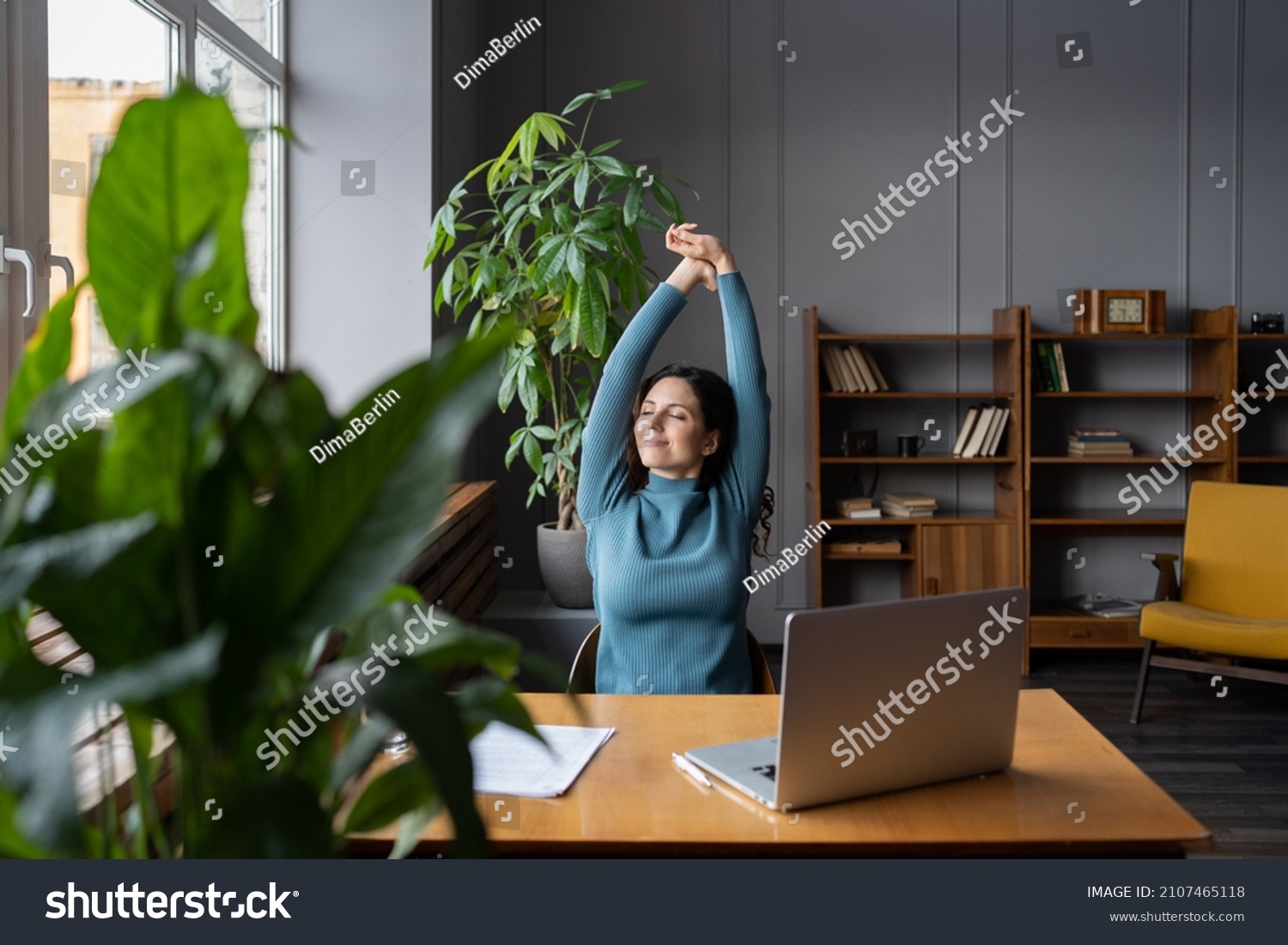 Relaxed office worker woman stretching hands and body taking break from work on laptop smiling look in window. Joyful freelancer copywriter girl happy with task done at workplace in coworking space #2107465118