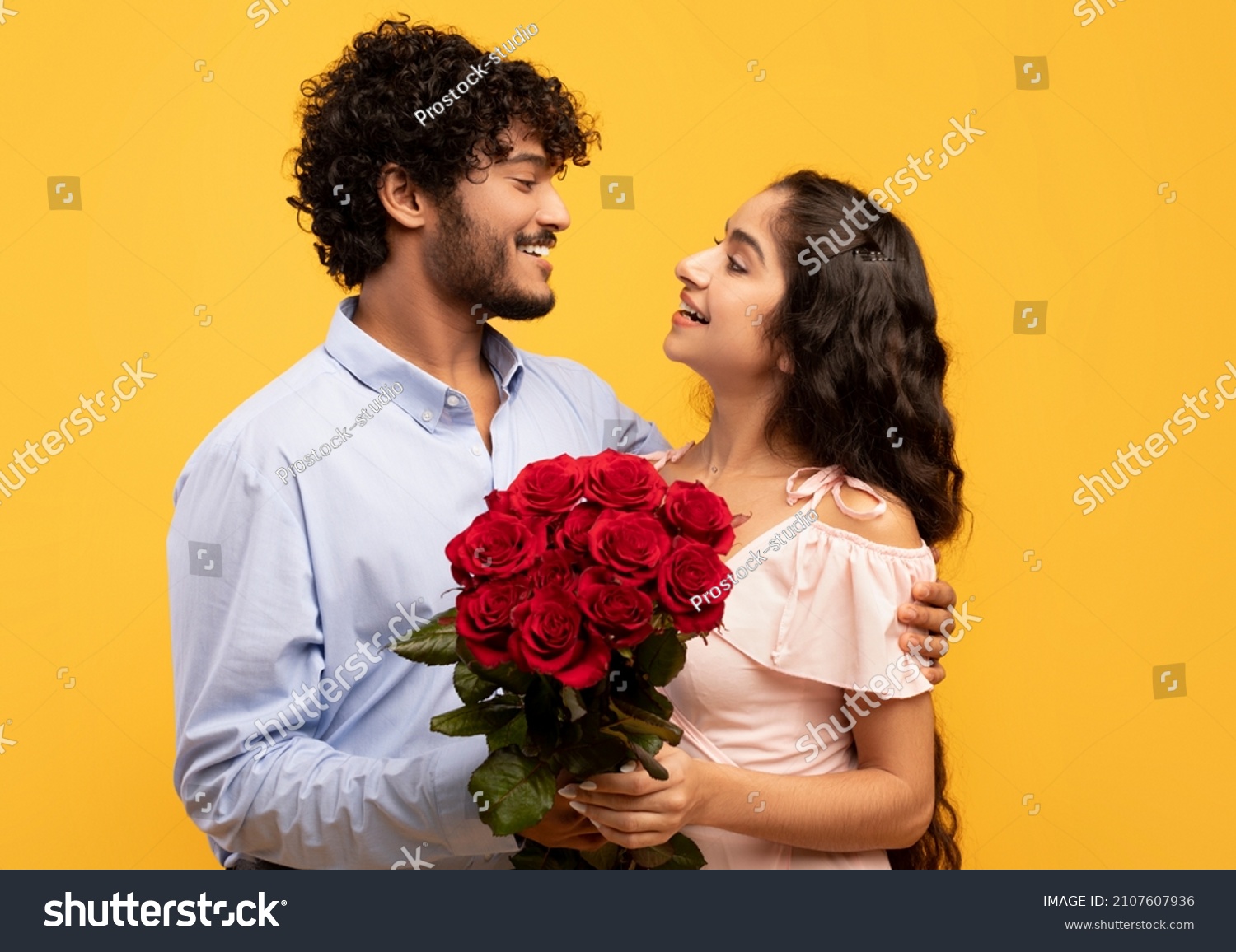 Lovers holiday. Loving indian couple holding bouquet of flowers, embracing and looking at each other on yellow background. Young guy pampering his girlfriend with roses on Valentine's Day #2107607936