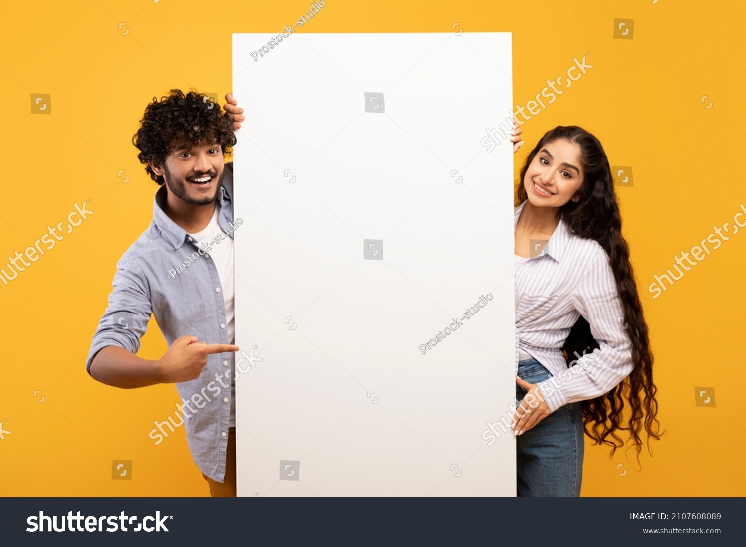Happy indian couple in love standing by white empty board for advertisement or text over yellow studio background. Excited man and woman standing next to blank placard for ad #2107608089