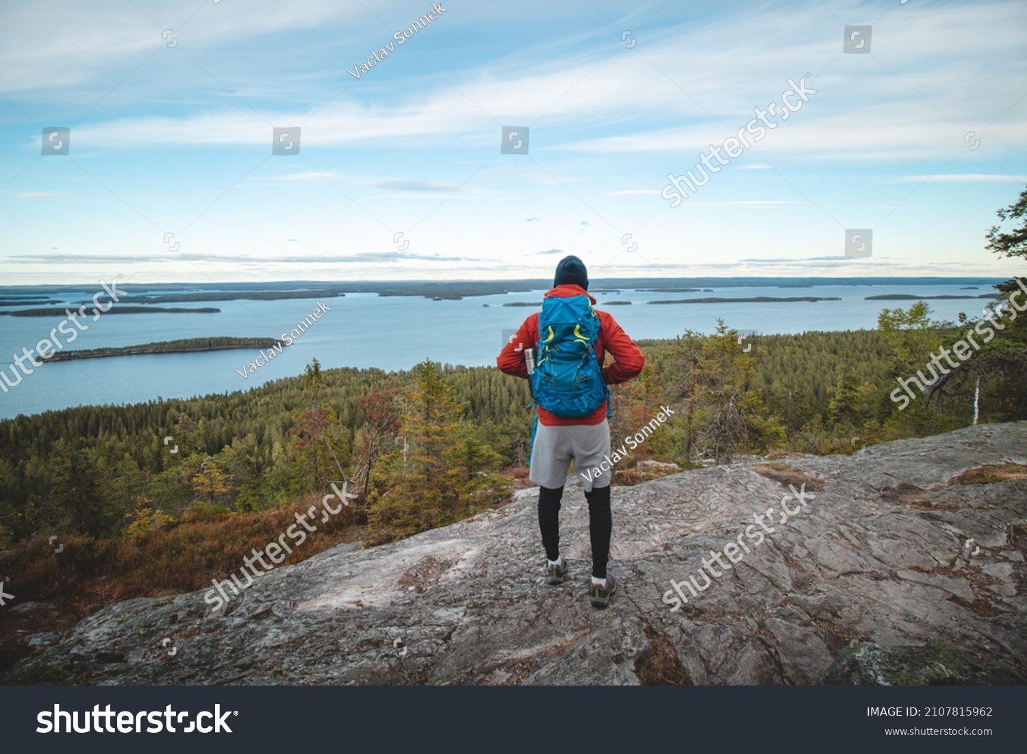 hiker wearing a jacket and carrying a backpack, standing on a rock watching Lake Jatkonjarvi at sunset in Koli National Park, eastern Finland. A man aged 24 wearing sports clothes. Active lifestyle. #2107815962