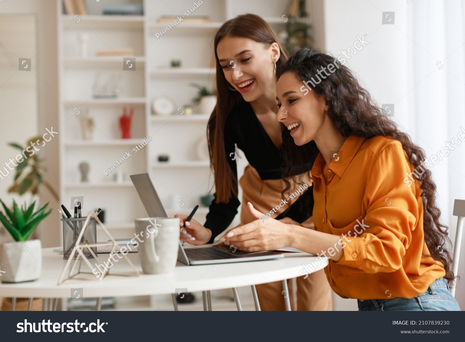 Two Happy Ladies Using Laptop Computer Working And Learning Online Together Or Making Video Call Sitting At Desk In Modern Office. E-Learning. Females Entrepreneurship Career Concept. Side View #2107839230